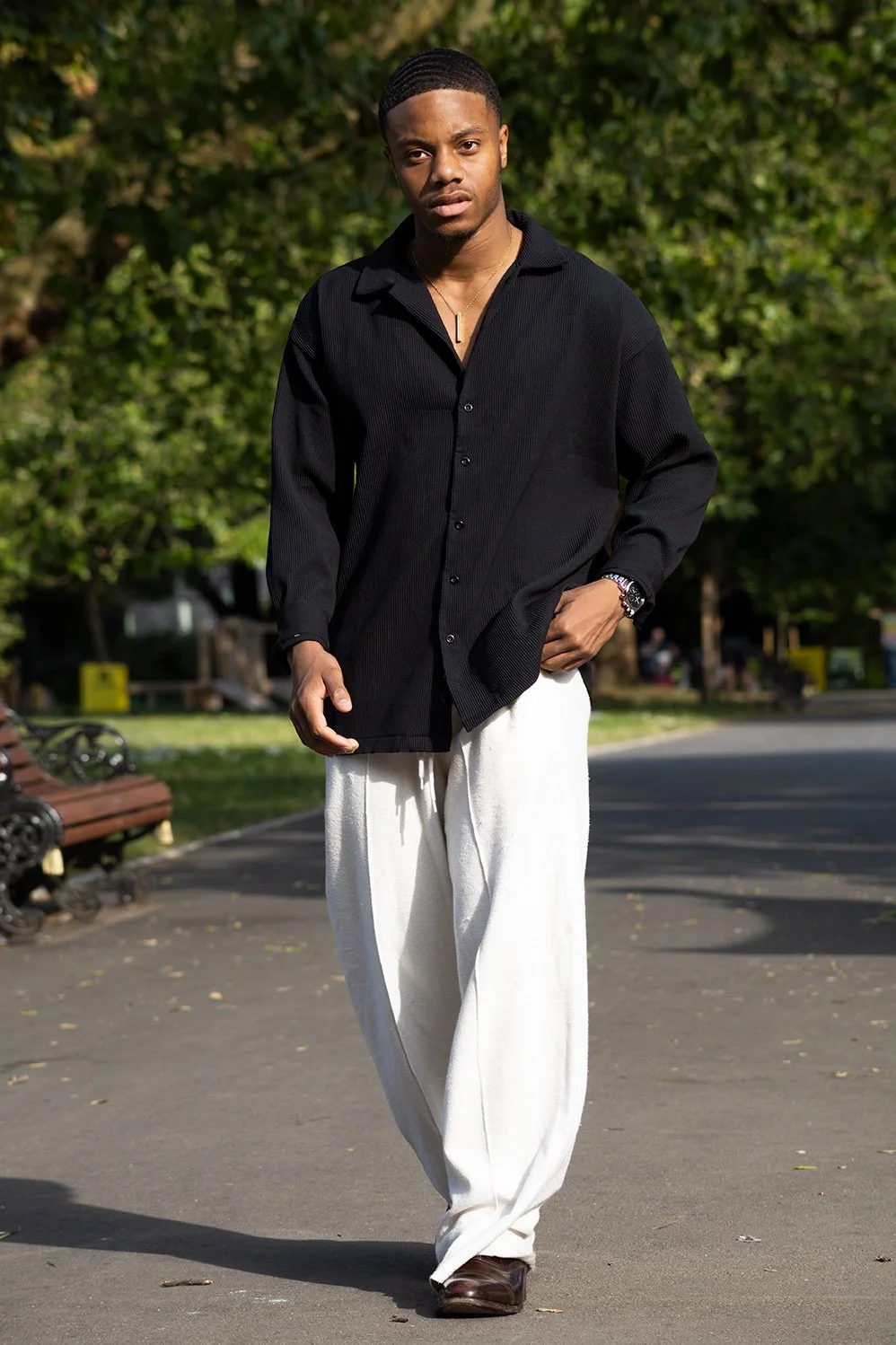 A young man walking outdoors in a park, wearing a black long-sleeve shirt, white pants, and dark shoes.