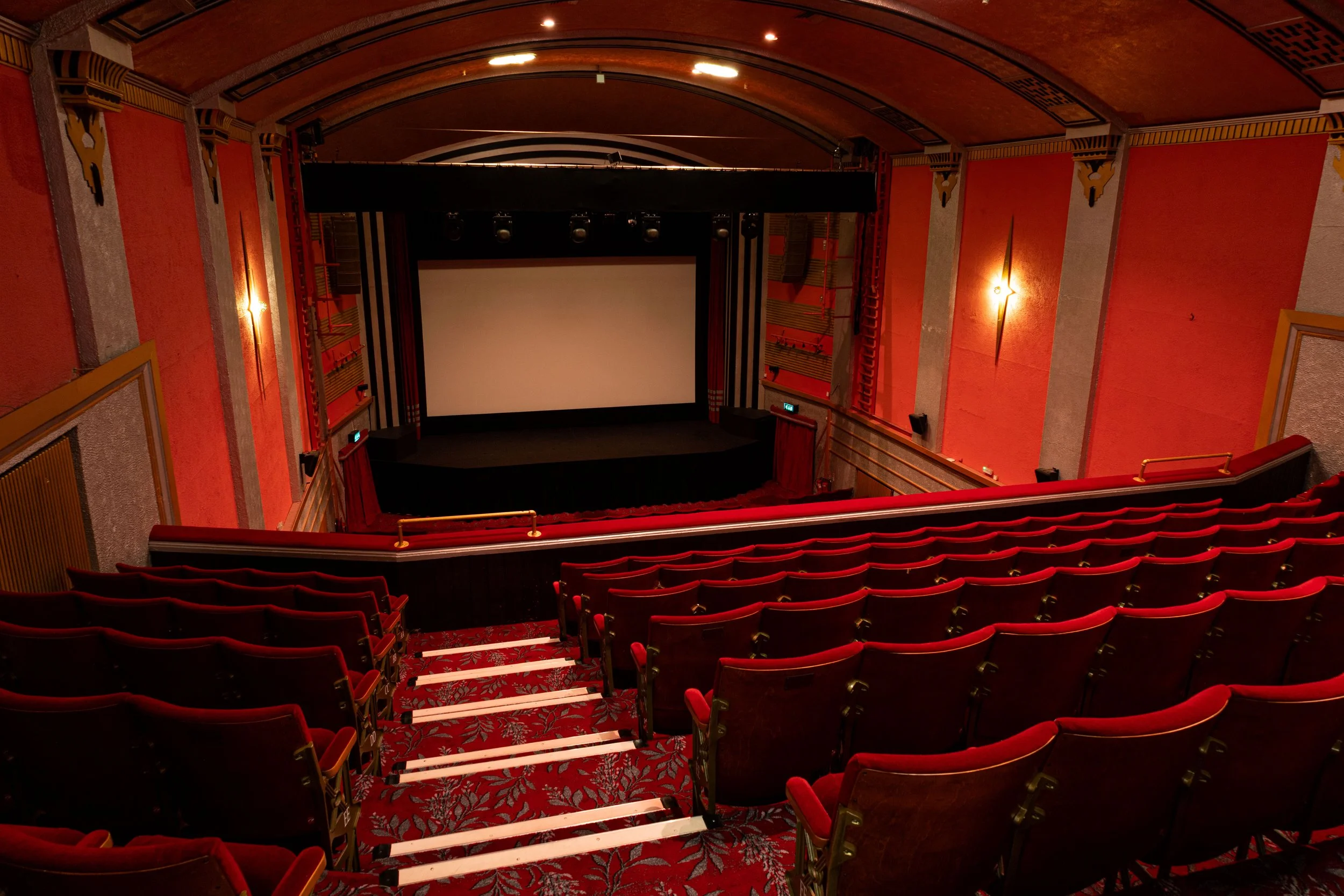 Photo of an empty theater auditorium with red chairs, a large screen, and a stage, viewed from the back rows.