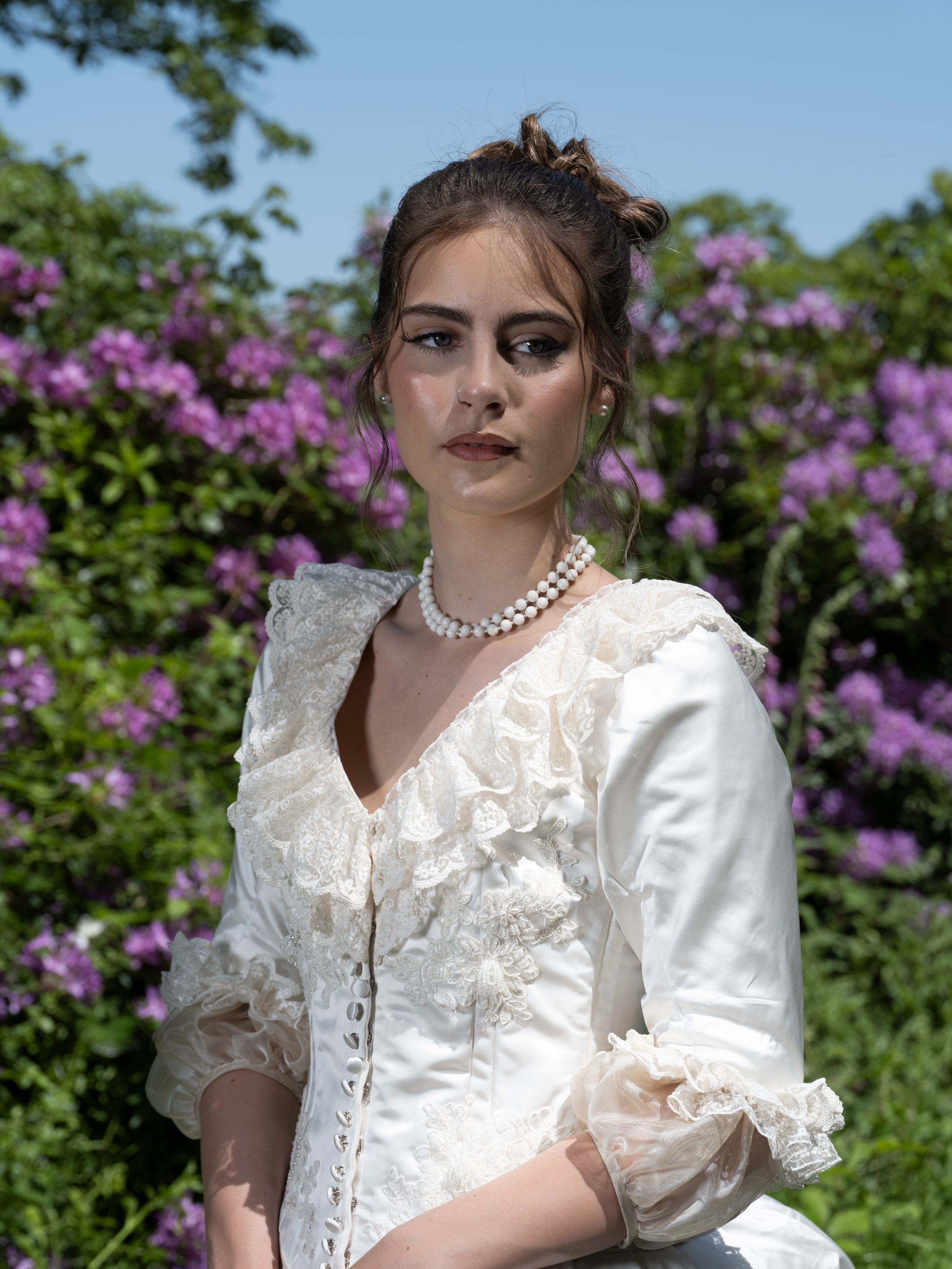 Portrait of a woman wearing a vintage white dress with lace details and pearl necklace, standing outdoors in front of purple flowering bushes and green foliage on a sunny day.