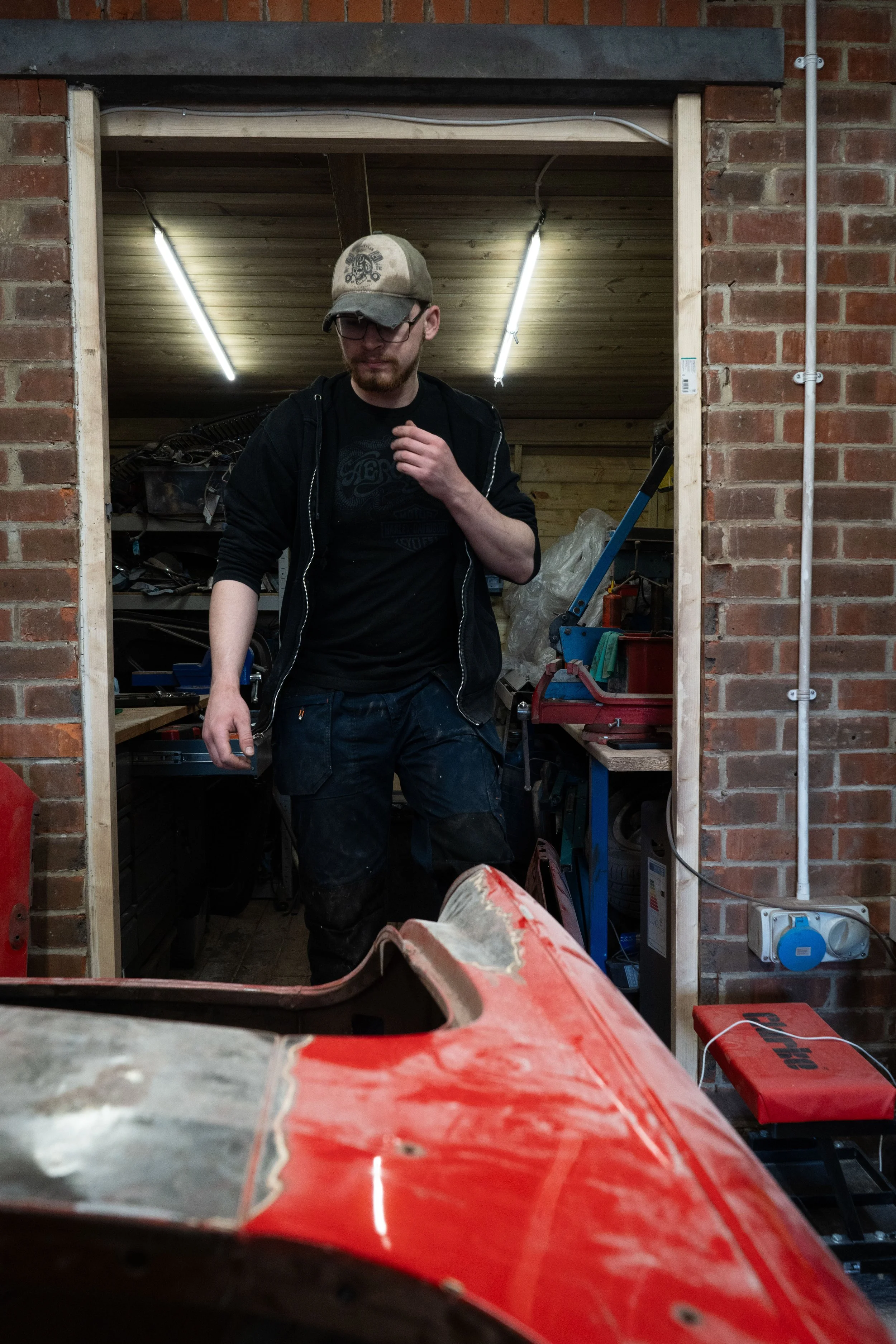 A man in a black hoodie, glasses, and a beige cap is standing in a workshop, with a brick wall behind him and tools and equipment around. He is looking down at something, with his right hand on his chest. The workshop has wooden ceiling panels, and t