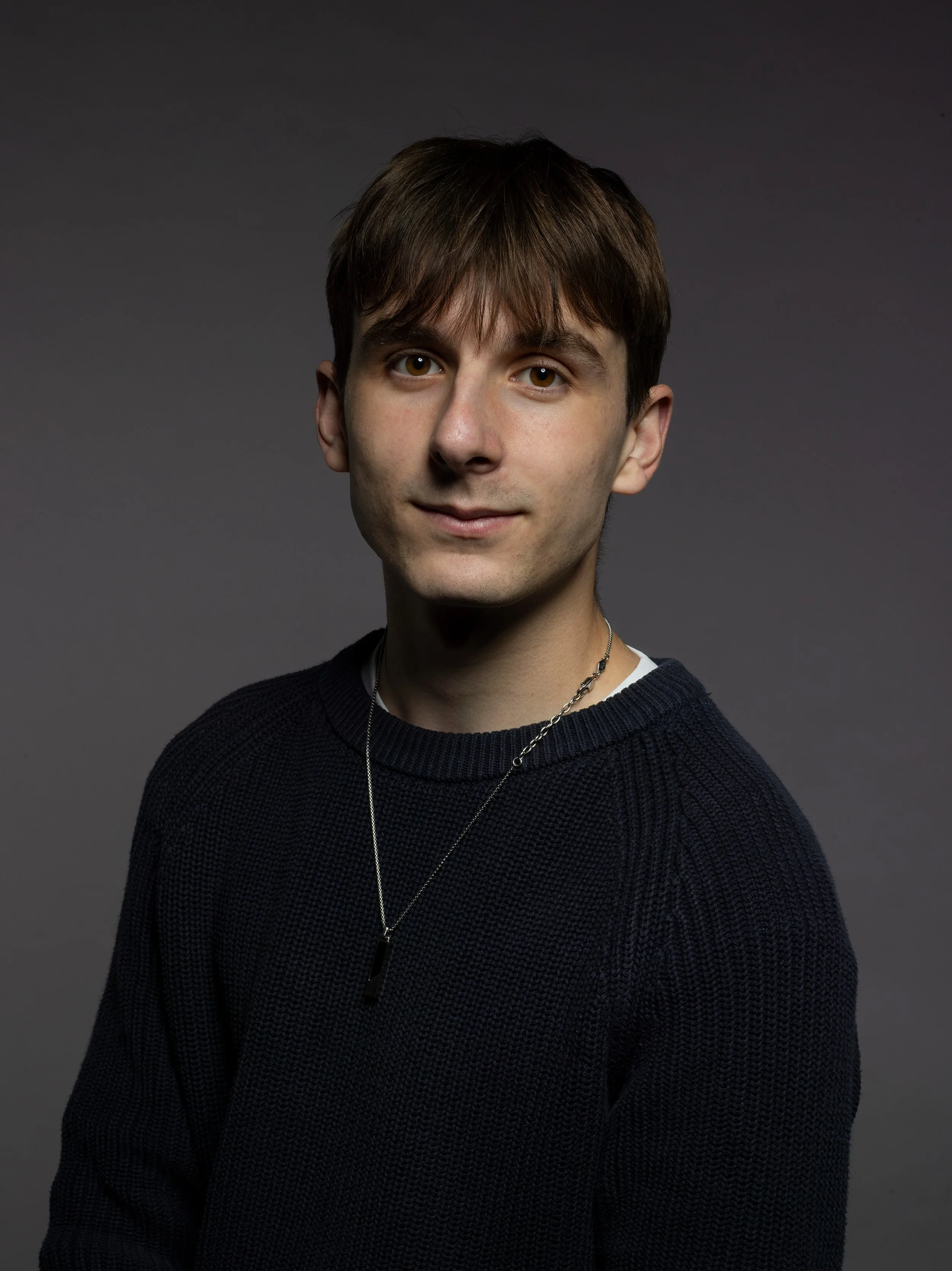 A young man with short dark brown hair, brown eyes, and fair skin wearing a black sweater and a layered necklace, posed against a dark gray background.
