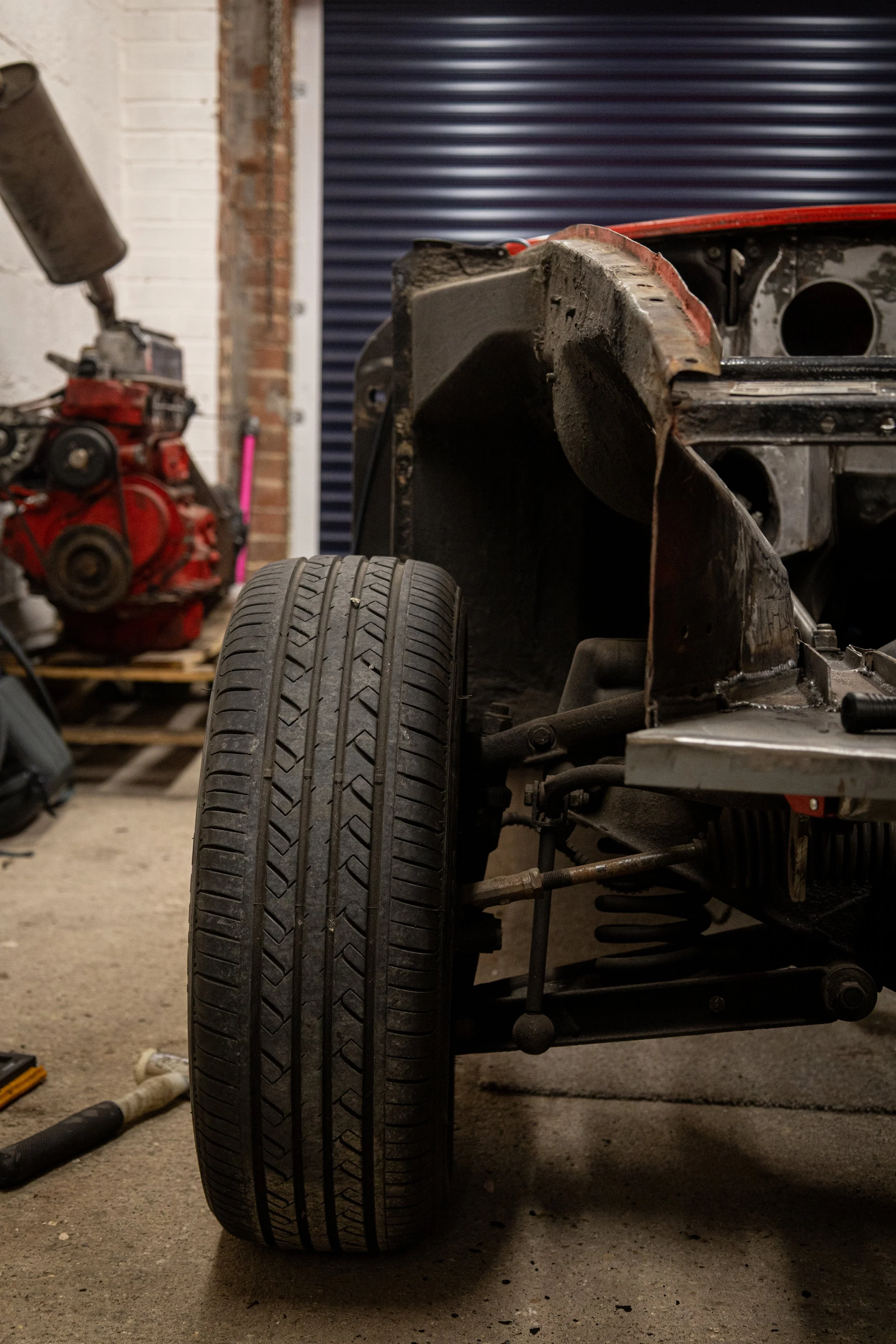 View of a partially disassembled vintage car showing front left tire, metal frame, and engine components in a garage with tools and equipment in the background.