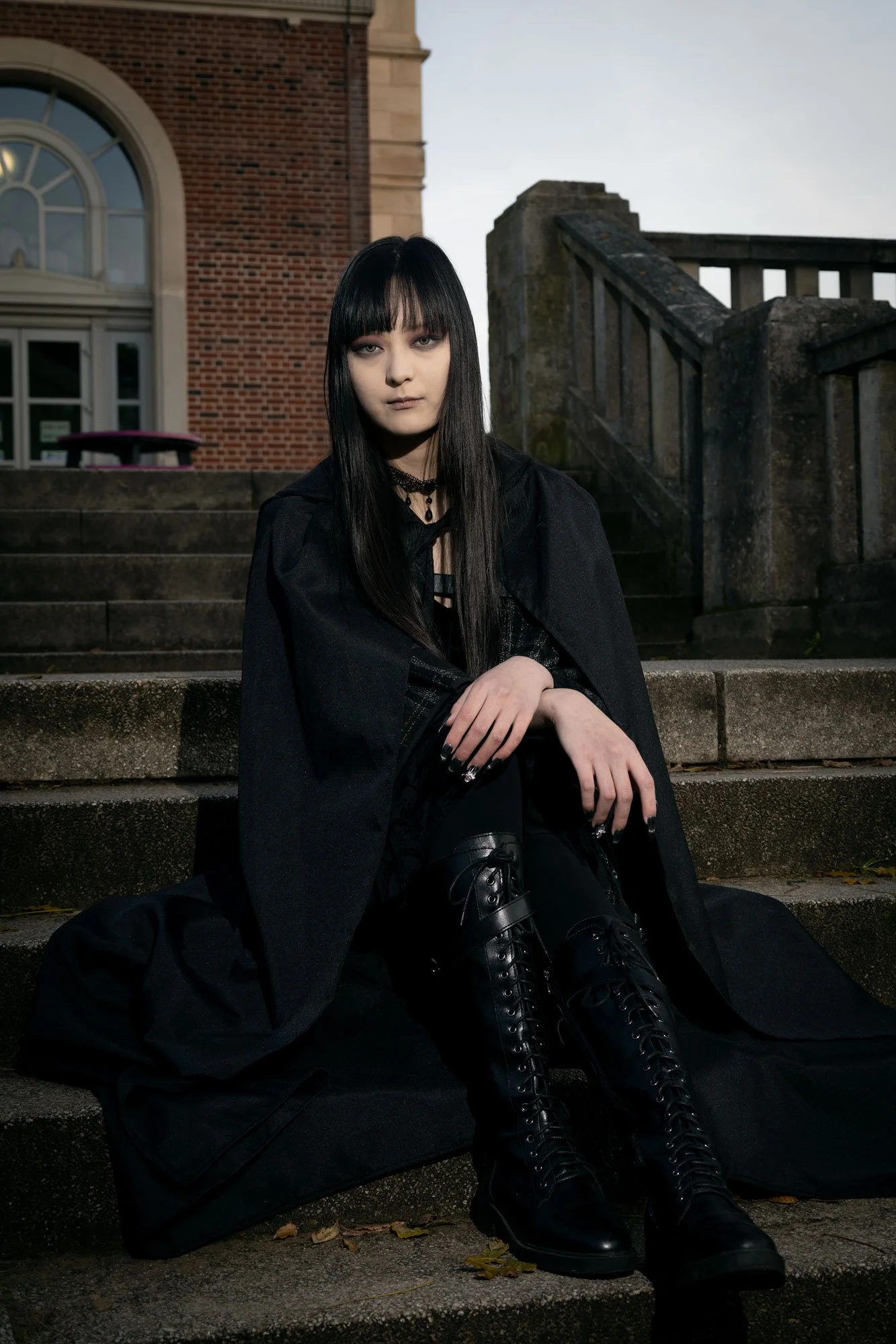 A young woman with long black hair and gothic fashion style sitting on stone steps outside a brick building with large arched windows. She is dressed in black with lace-up boots, a choker, and dark clothing, with a black bag beside her.