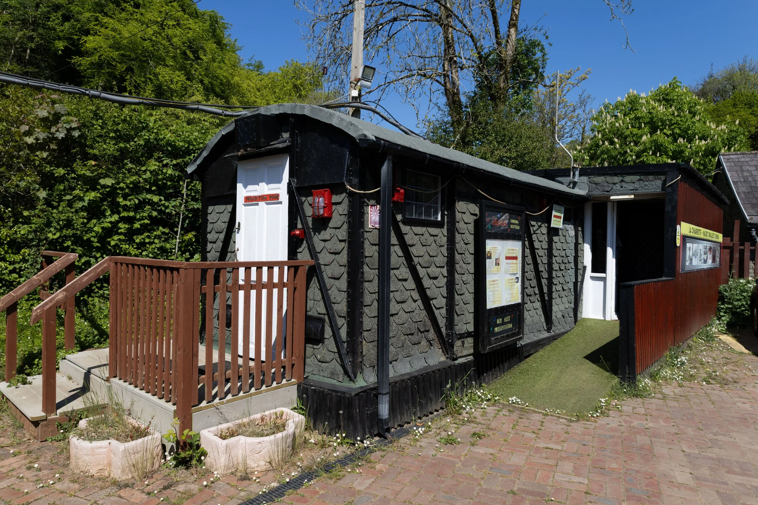 Small building with shingle siding, black upper walls, and a white door labeled 'Projection Room'. There is a red fire alarm and a red fire extinguisher mounted outside. Posters are displayed on the exterior wall, with a small wooden ramp and planter