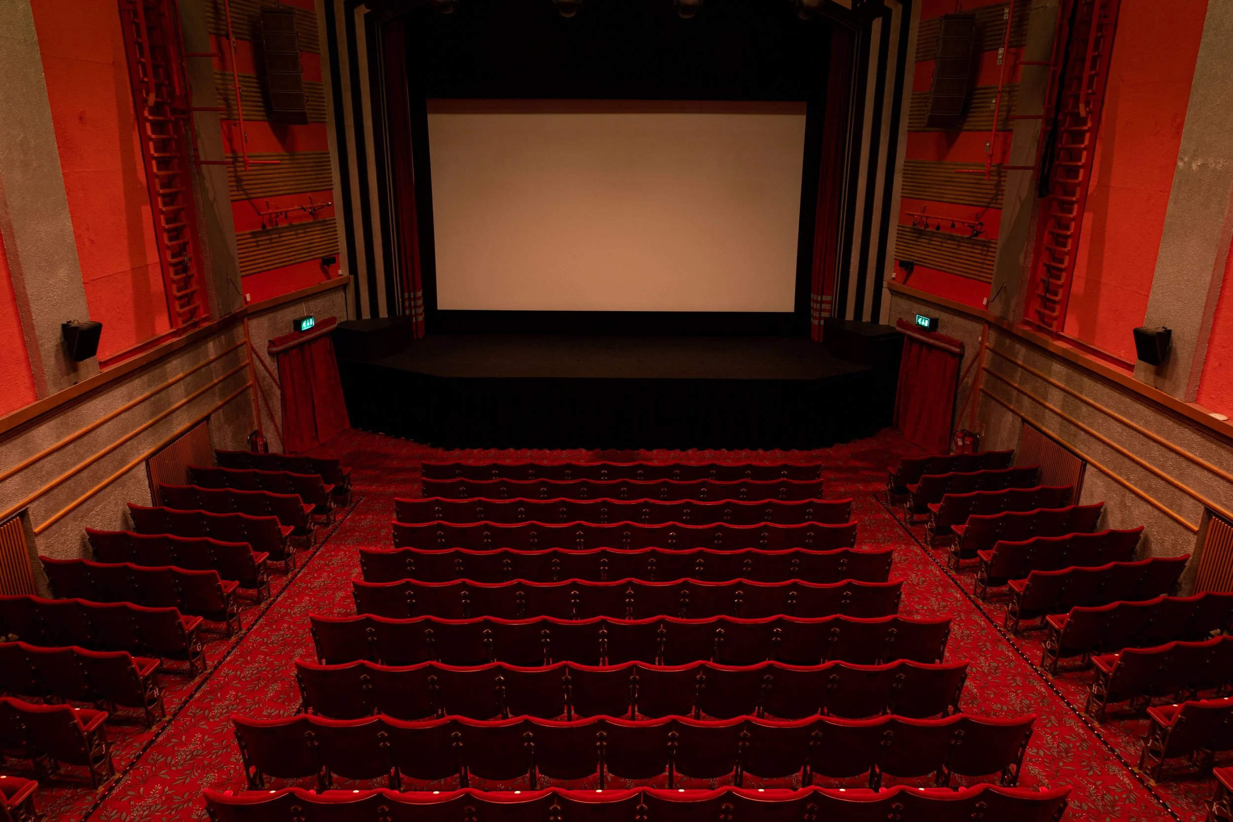 This is an image of a movie theater auditorium with rows of red seats facing a large empty movie screen. The walls are decorated with red and beige patterns.