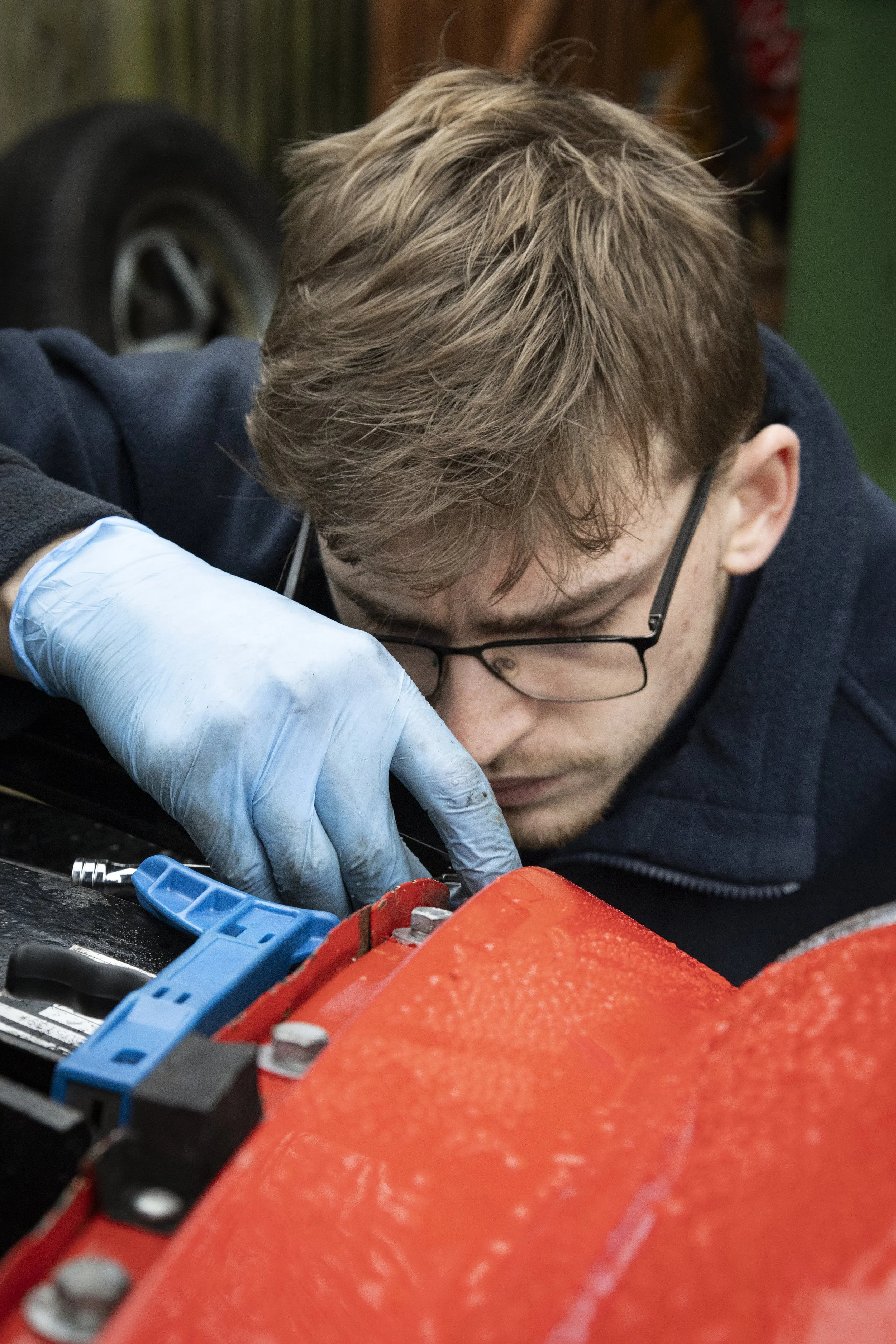 A person with glasses and light brown hair working on a mechanical project, wearing blue gloves and focused on the task.