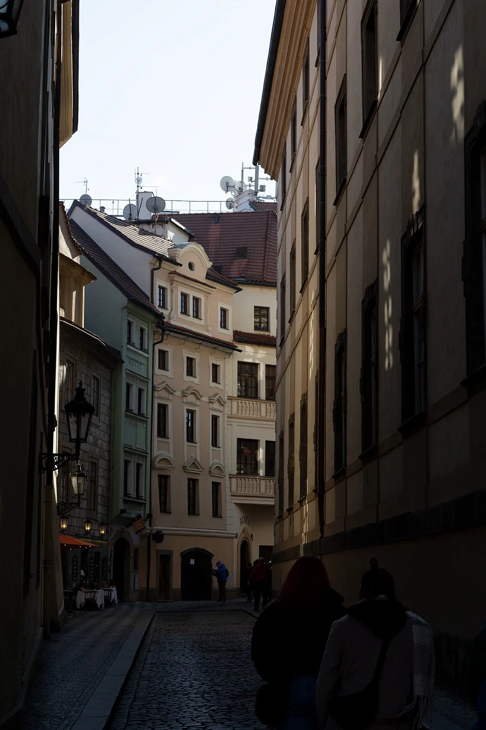 Narrow cobblestone alleyway in a European city with colorful historic buildings, a few people walking, and rooftops with satellite dishes visible.