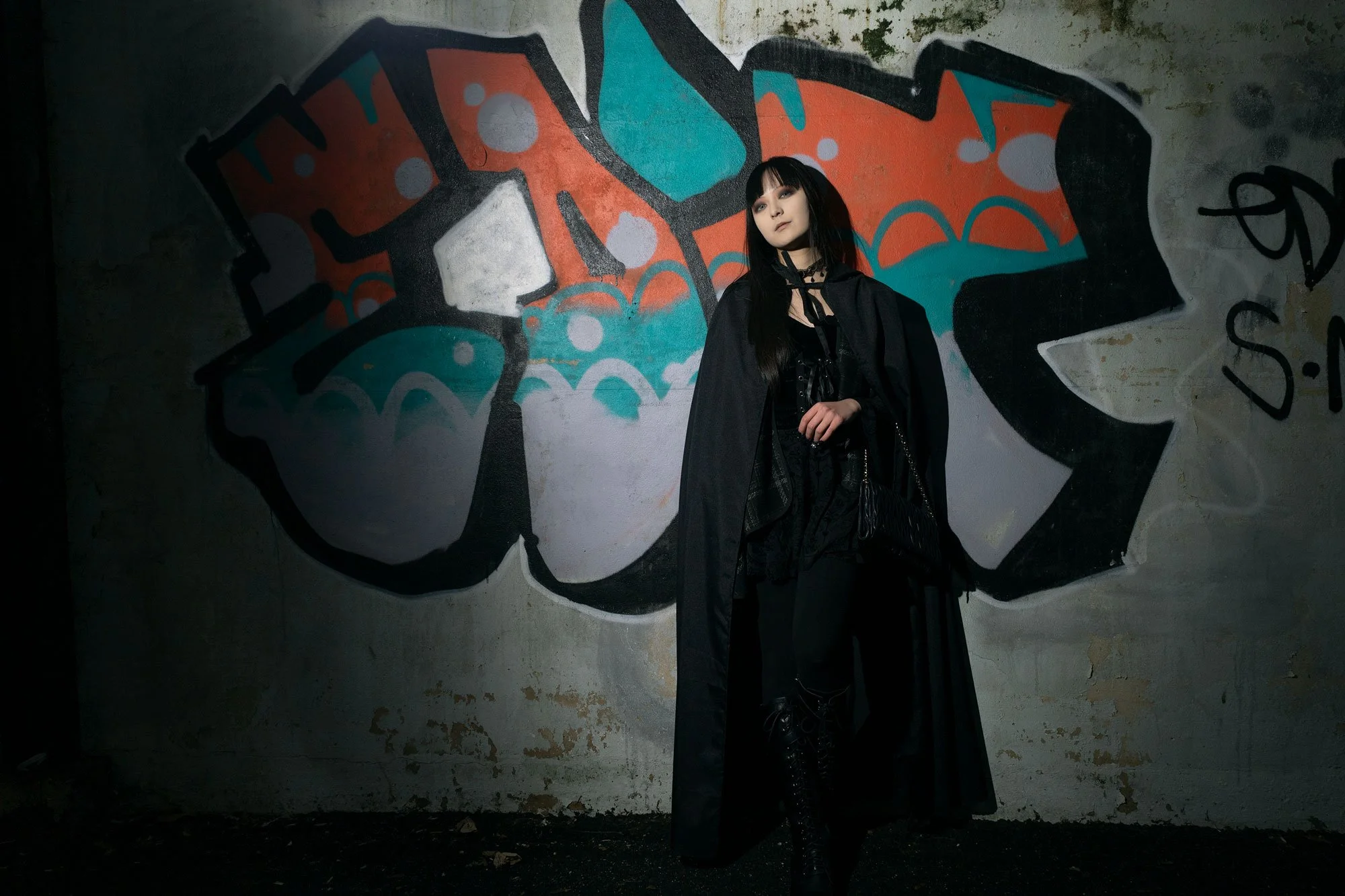 A young woman dressed in black, with long black hair and gothic fashion, standing in front of a colorful graffiti mural on a concrete wall in an urban setting.