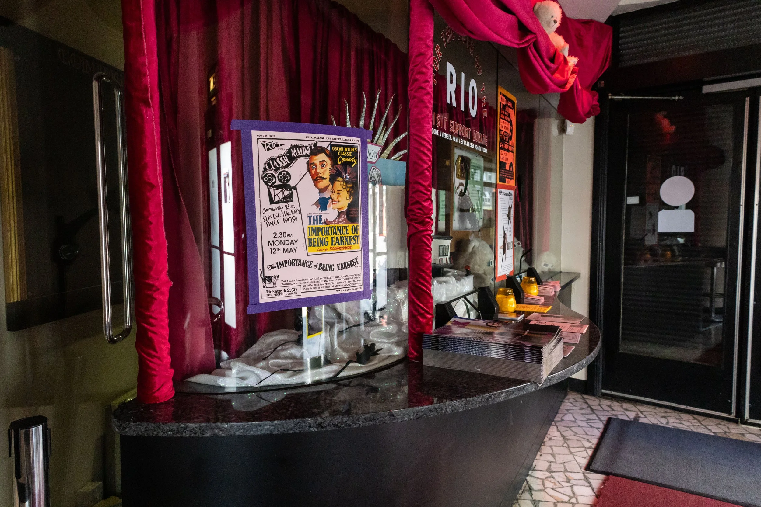 Indoor scene of a reception or information desk with posters. One poster promotes a comedy titled 'The Importance of Being Earnest' at Rio Cinema. Red curtains frame the desk, with pamphlets and small yellow lights on the counter.