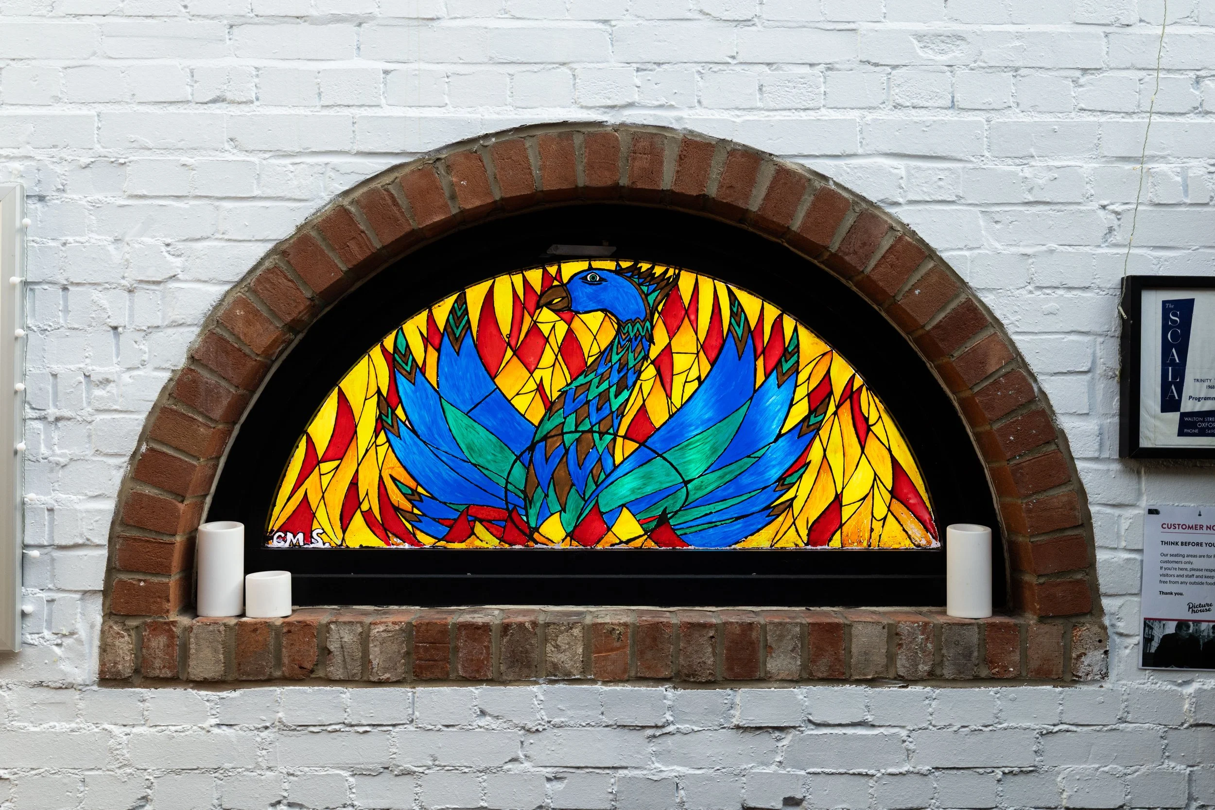 Colorful stained glass window depicting a peacock with vibrant blue, green, yellow, and red feathers, set in a brick archway against a white brick wall.