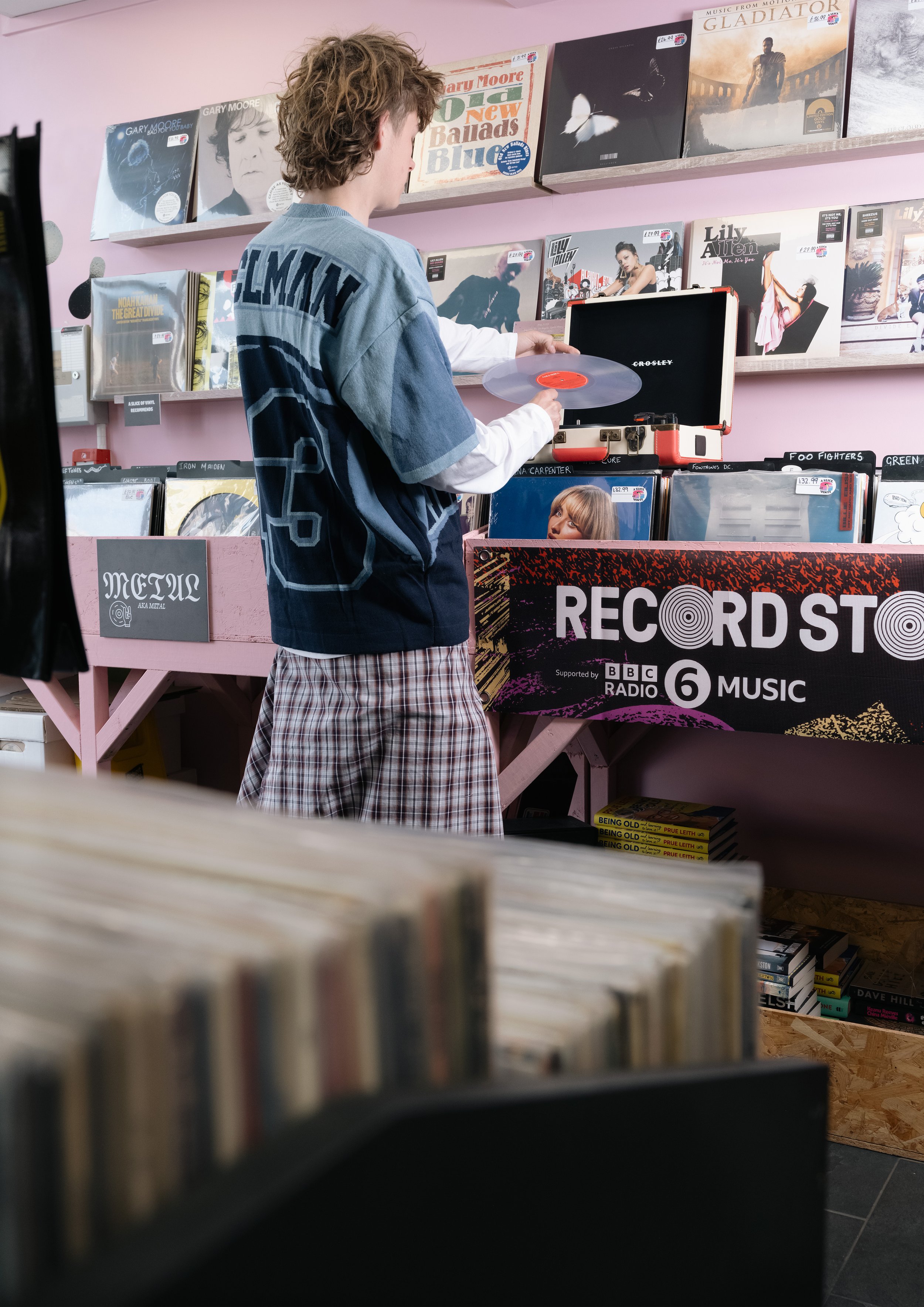 A person with curly hair wearing a graphic t-shirt and plaid shorts browsing vinyl records in a record store with pink walls. They are holding a vinyl record over an open record player.