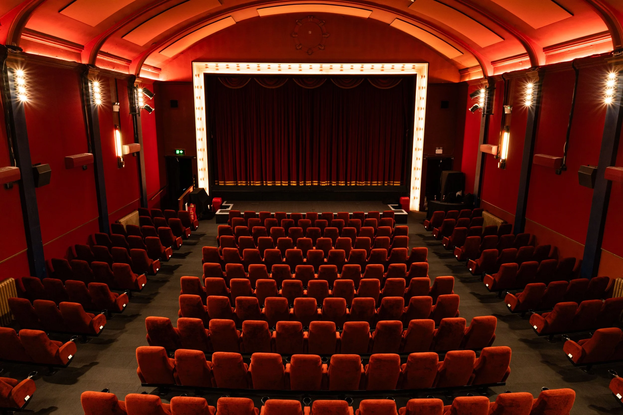 Empty theater with a stage, red velvet curtains, red upholstered seats, and warm ambient lighting.