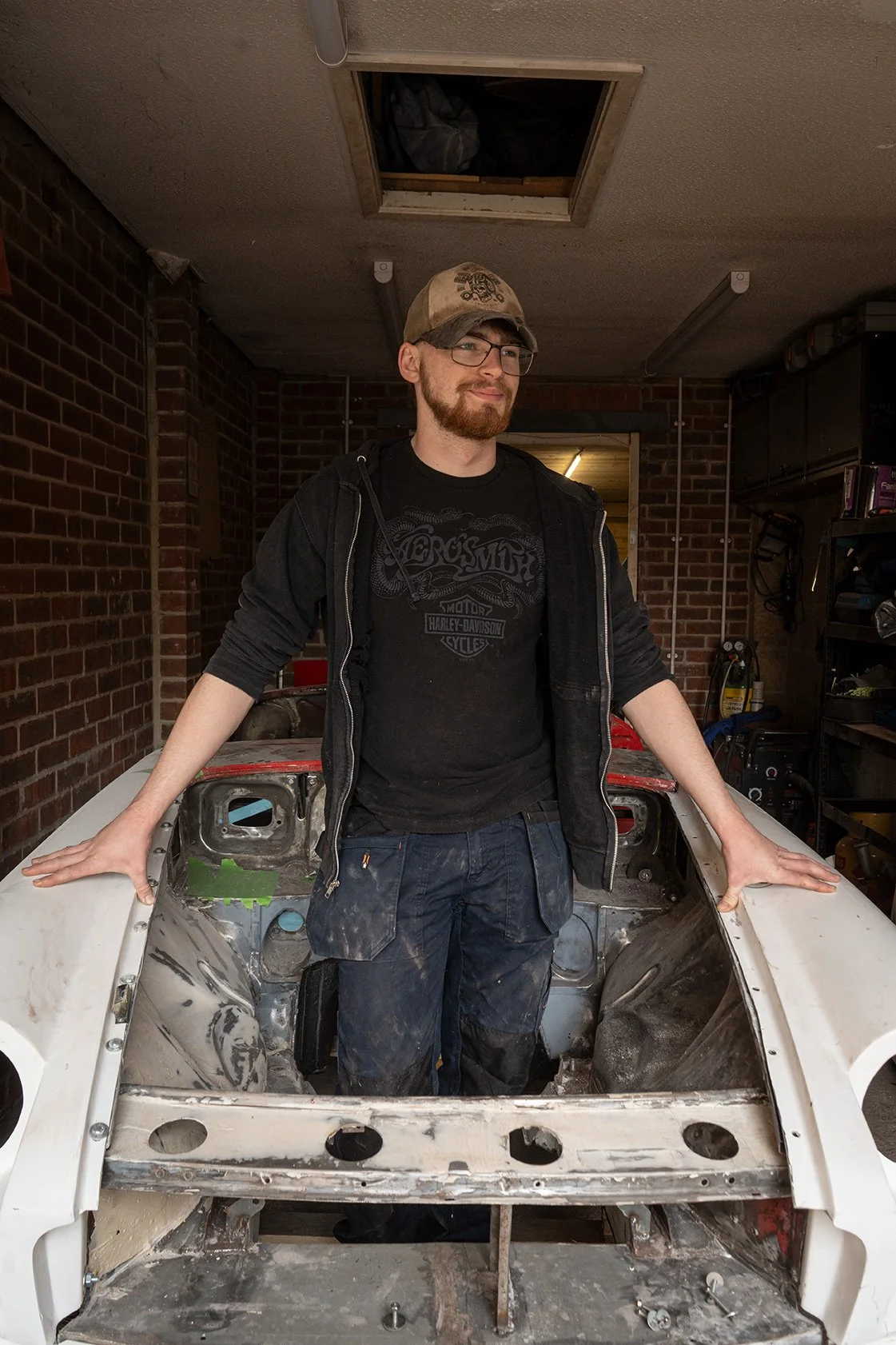 A man with a beard, glasses, wearing a cap, black hoodie, and black T-shirt standing inside a car frame in a garage. The garage has brick walls and shelves with tools.