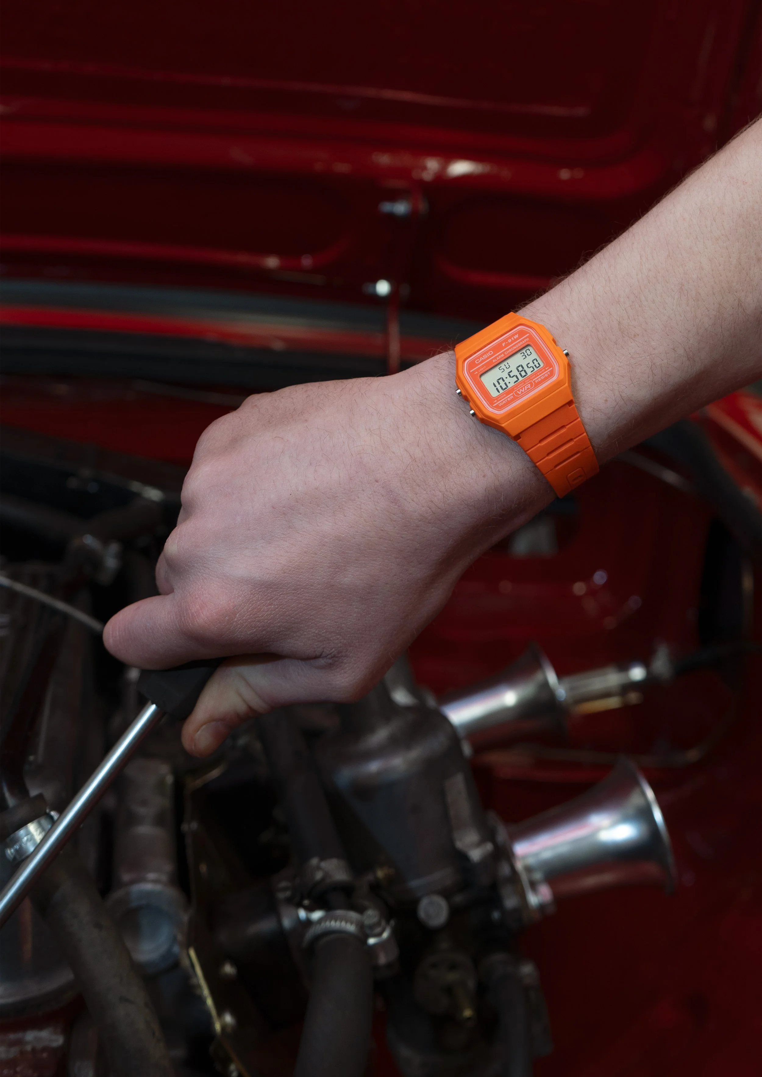 A person's arm wearing an orange digital watch, holding a screwdriver, with a background of machinery and red-painted metal pipes.