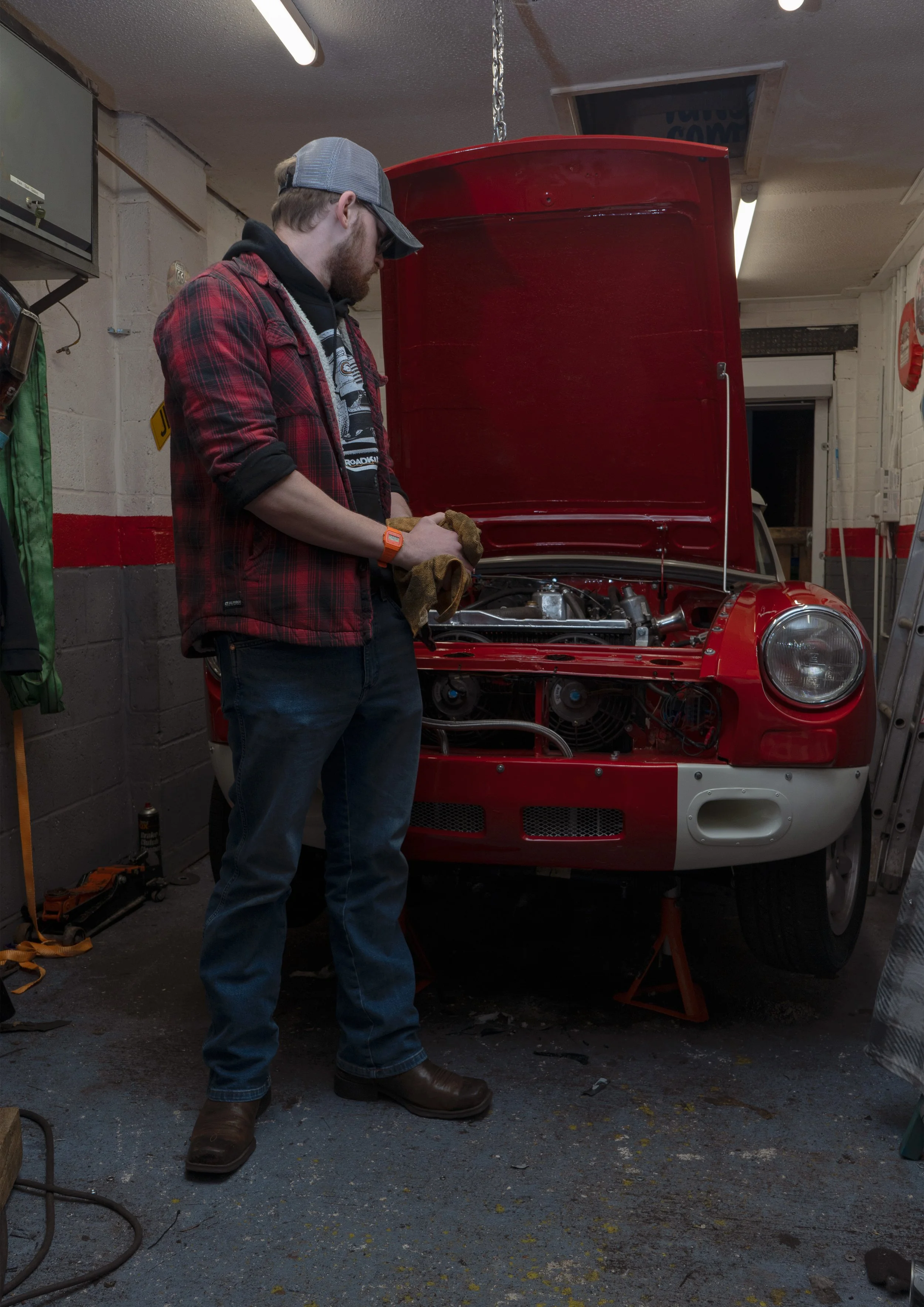 A man wearing a gray cap, sunglasses, a red and black plaid shirt, dark jeans, and brown boots is working on a red race car with its hood open in a garage. The garage has a concrete wall with a green coat hanging on hooks, and various tools and equip