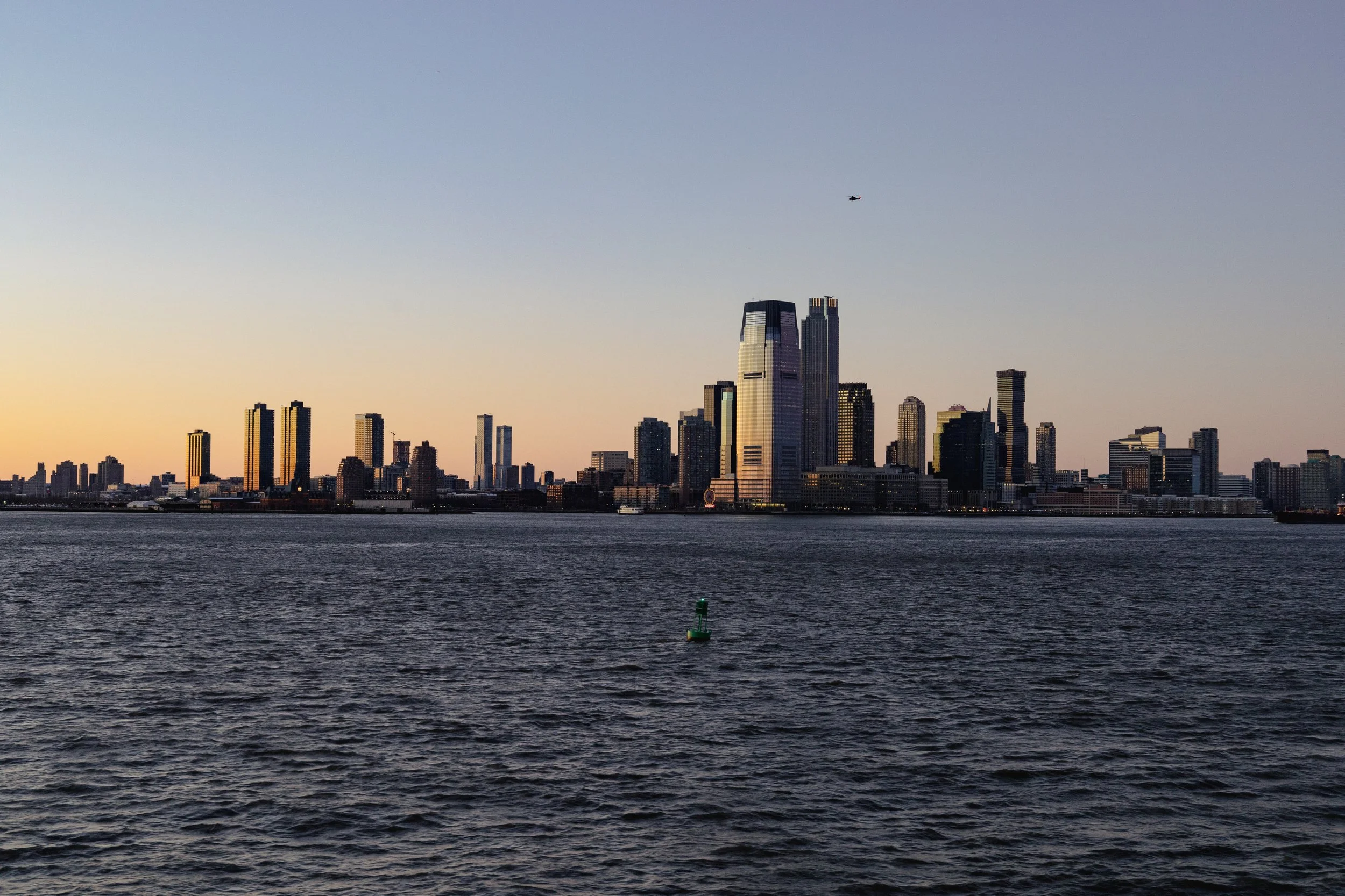 Skyline of a city with tall buildings along a water body during dusk, with a helicopter in the sky.