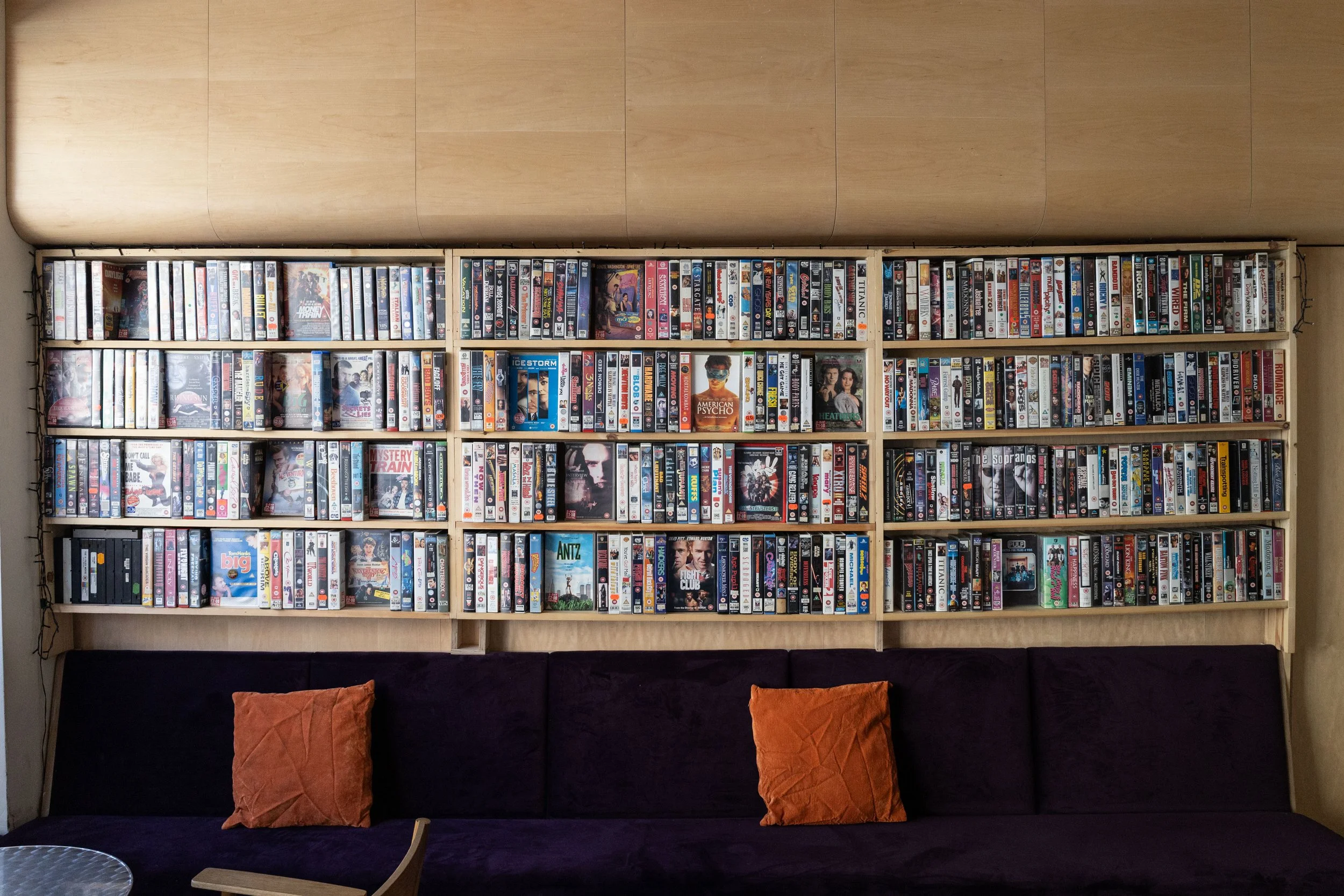 A large wooden bookshelf filled with DVDs arranged in rows, placed against a wooden wall. In front of the bookshelf is a black couch with two orange pillows and a small wooden table.