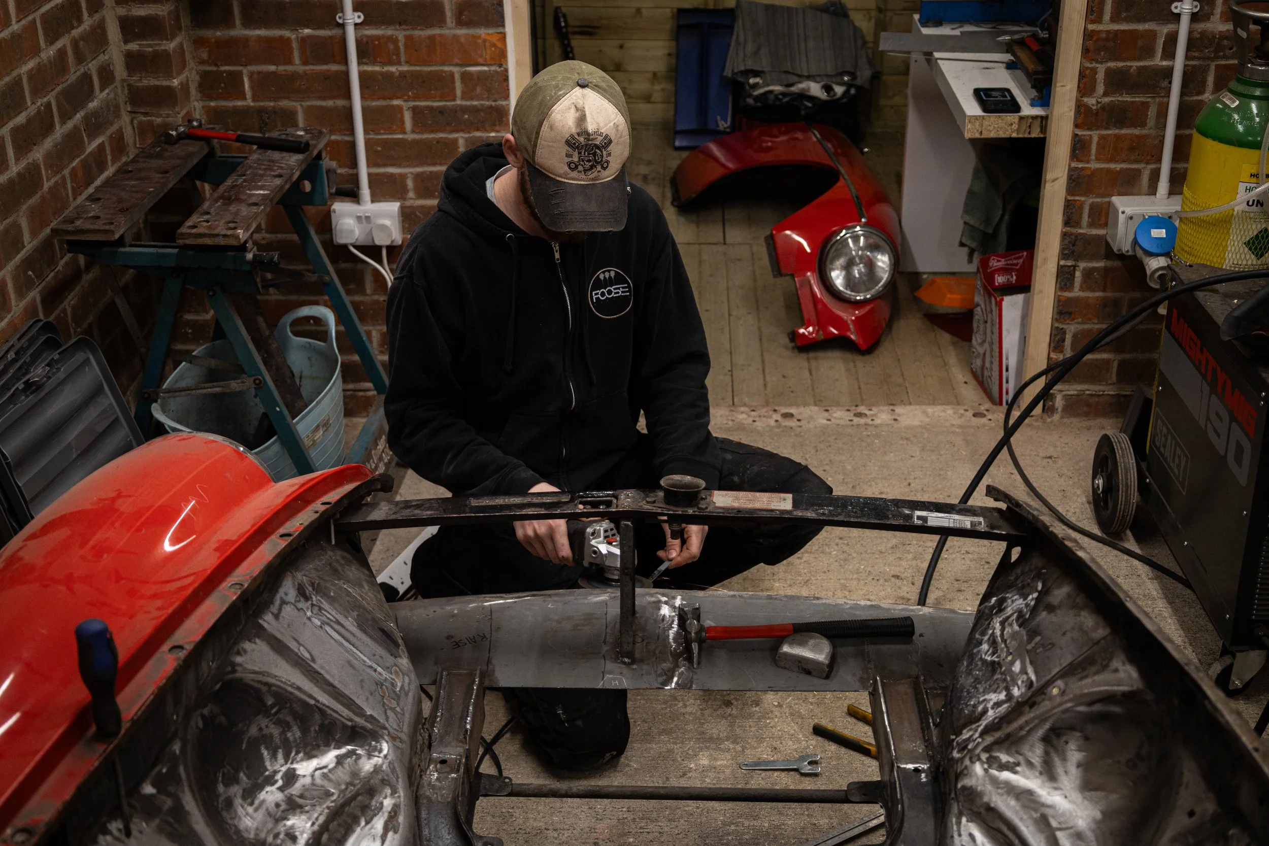 A person working on a small vehicle frame in a garage. The person is wearing a black hoodie and a beige cap. The garage has brick walls, tools, and equipment, including an orange motorcycle or scooter with a partially assembled frame.