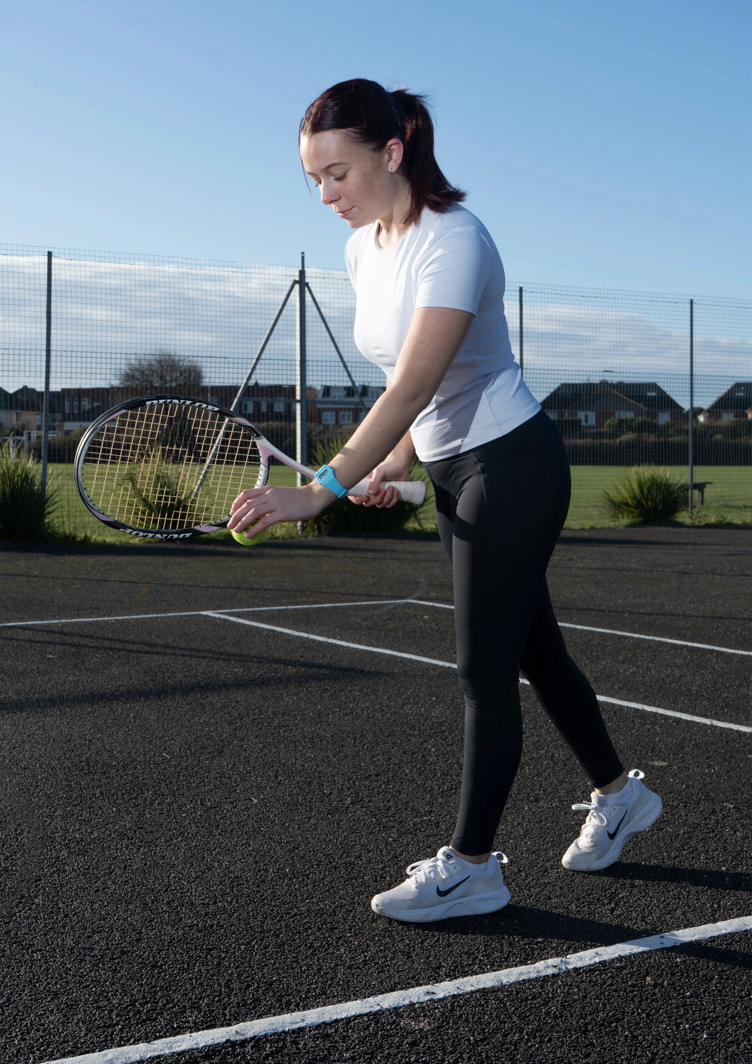 A woman playing tennis on an outdoor court, holding a tennis racket and preparing to serve, with houses and a fence in the background on a clear day.