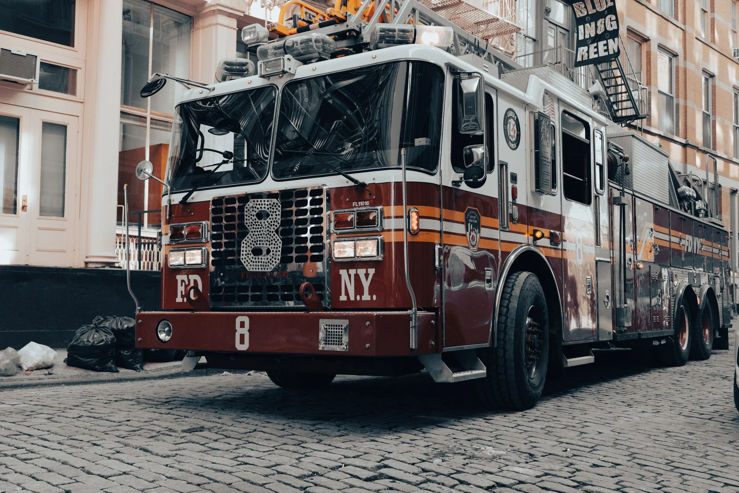 A fire truck with ladder equipment parked on a brick street in front of a building with brick and cream-colored walls. The truck has a large number 8 on the front and various firefighting tools on top.