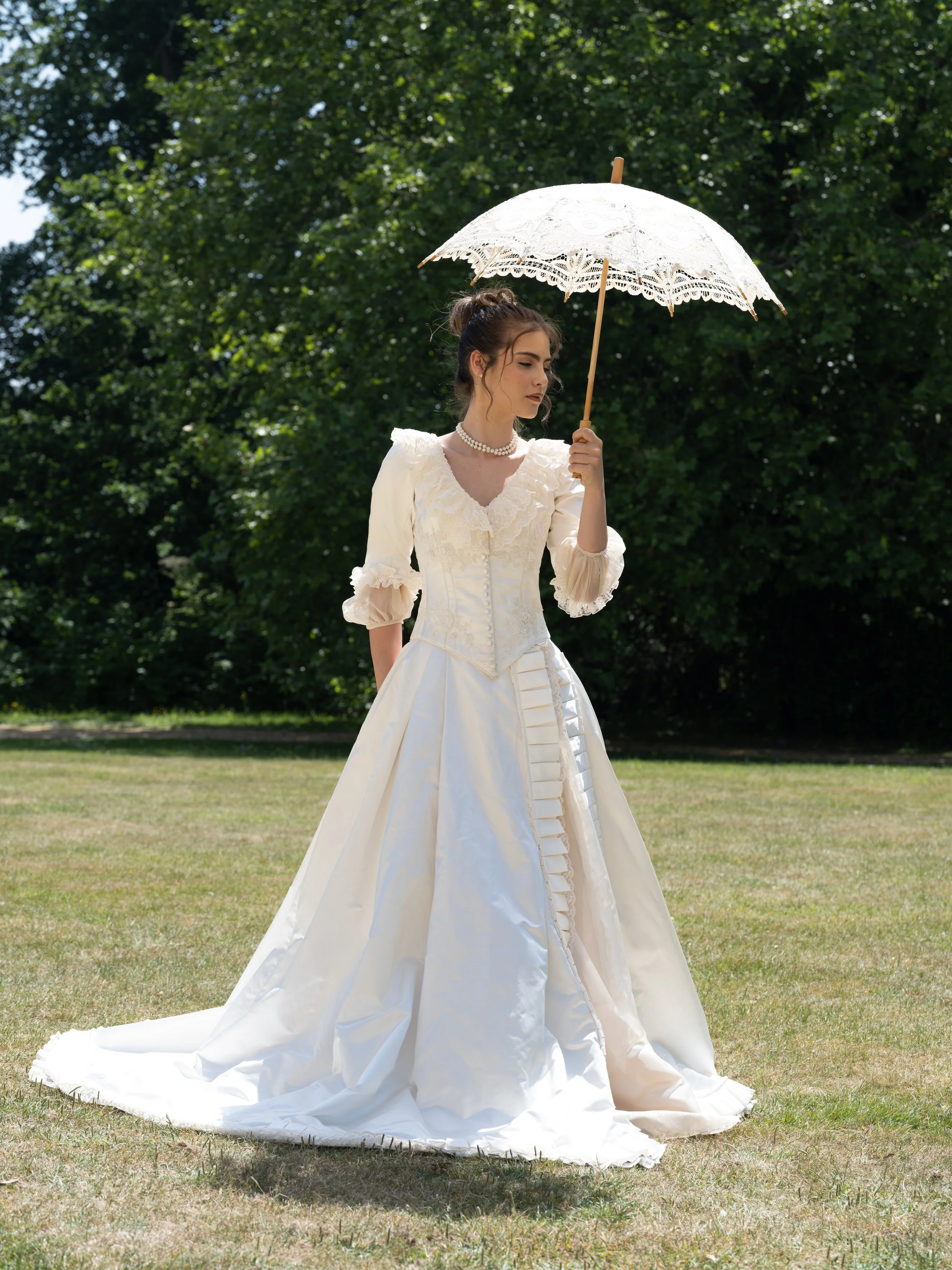 Young woman wearing a vintage white wedding dress holding a lace parasol outdoors on a sunny day, with trees in the background.