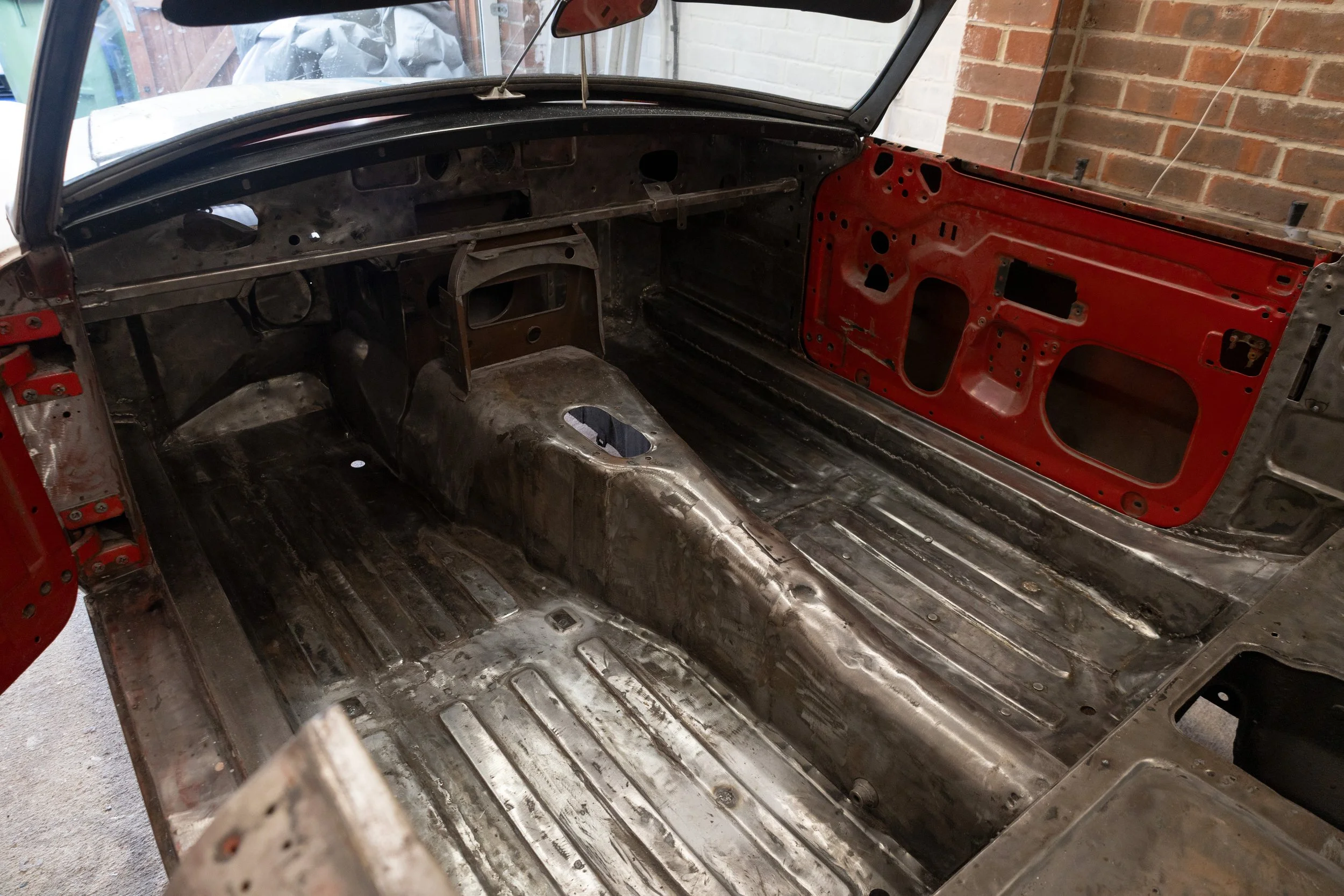 Interior of a car stripped down to metal, showing the dashboard area and the floor with no seats or interior panels, with a brick wall background.