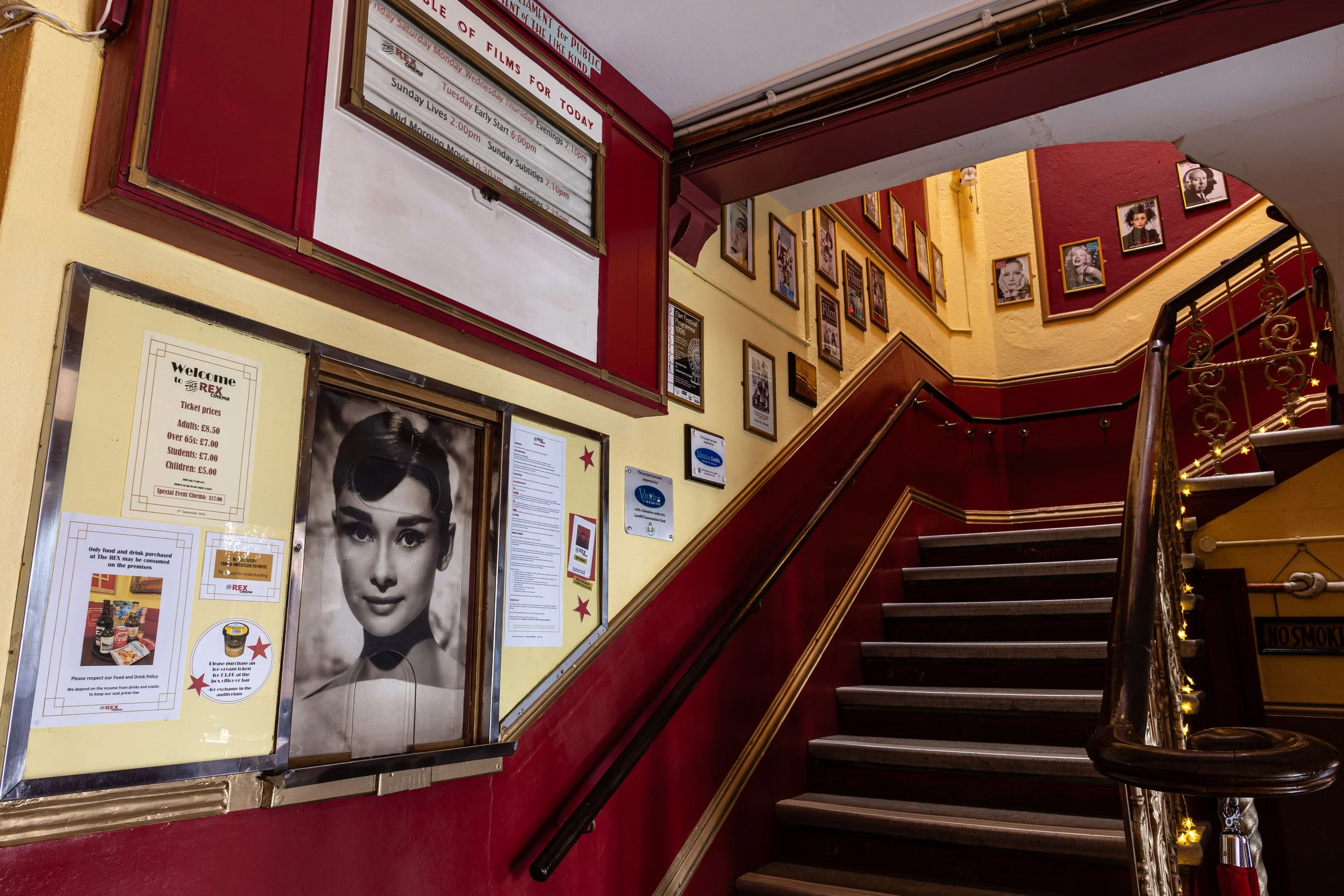 Interior of a theater or cinema lobby with a staircase, framed photos, and a schedule display. The walls are painted red and yellow, with framed portraits of classic movie stars. There are also informational posters and ticket prices displayed.