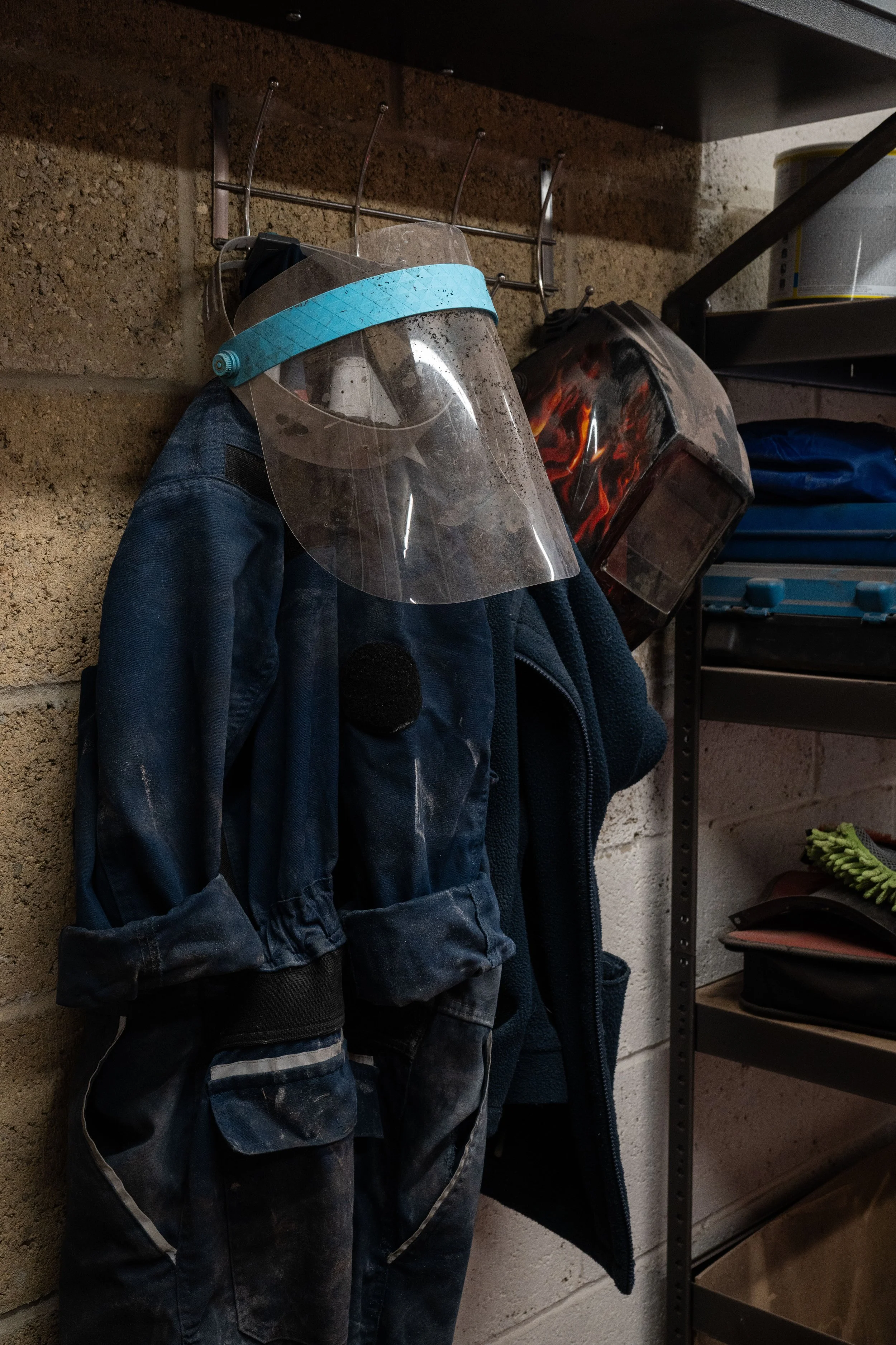 A collection of safety gear hanging on a wall including a clear face shield with a blue strap, a black and red welding helmet with flames design, and a dark jacket. Shelves in the background hold containers and other miscellaneous items.