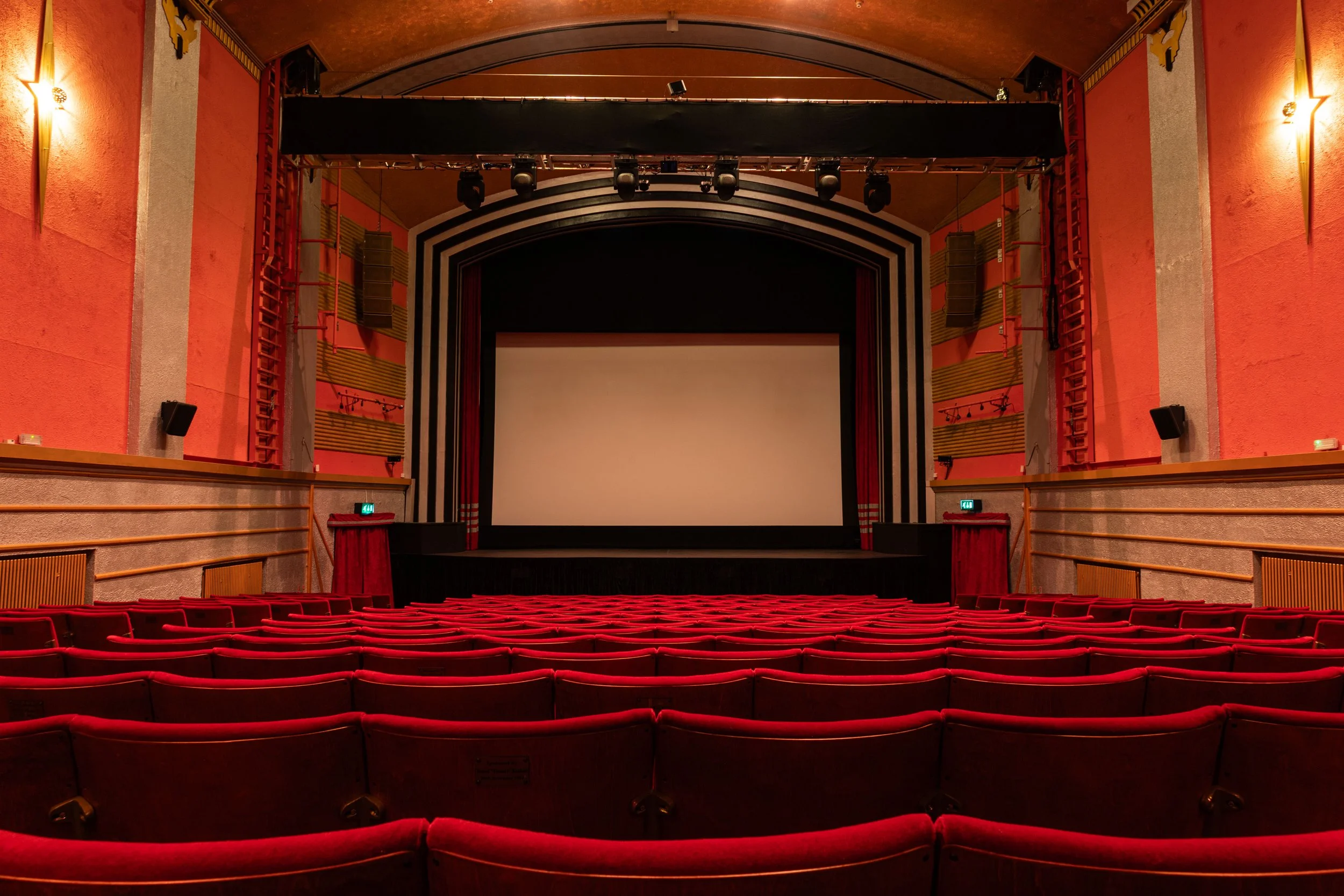 Inside view of a theater or cinema with rows of red seats facing a large stage with a blank white screen, surrounded by red curtains and black and white striped framing, with stage lights and speakers.
