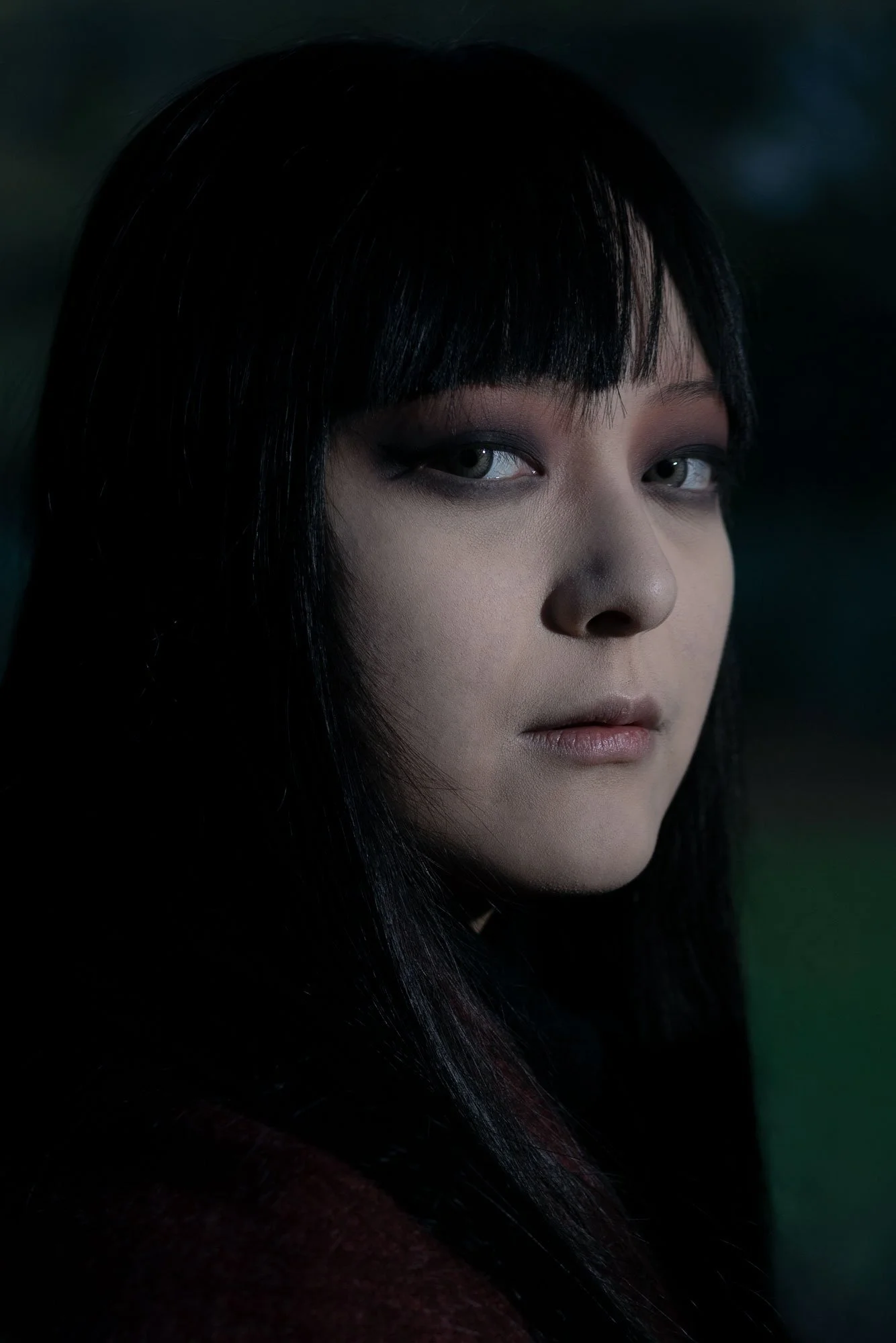 Close-up portrait of a woman with black hair and light skin, looking over her shoulder with a serious expression, dark background.