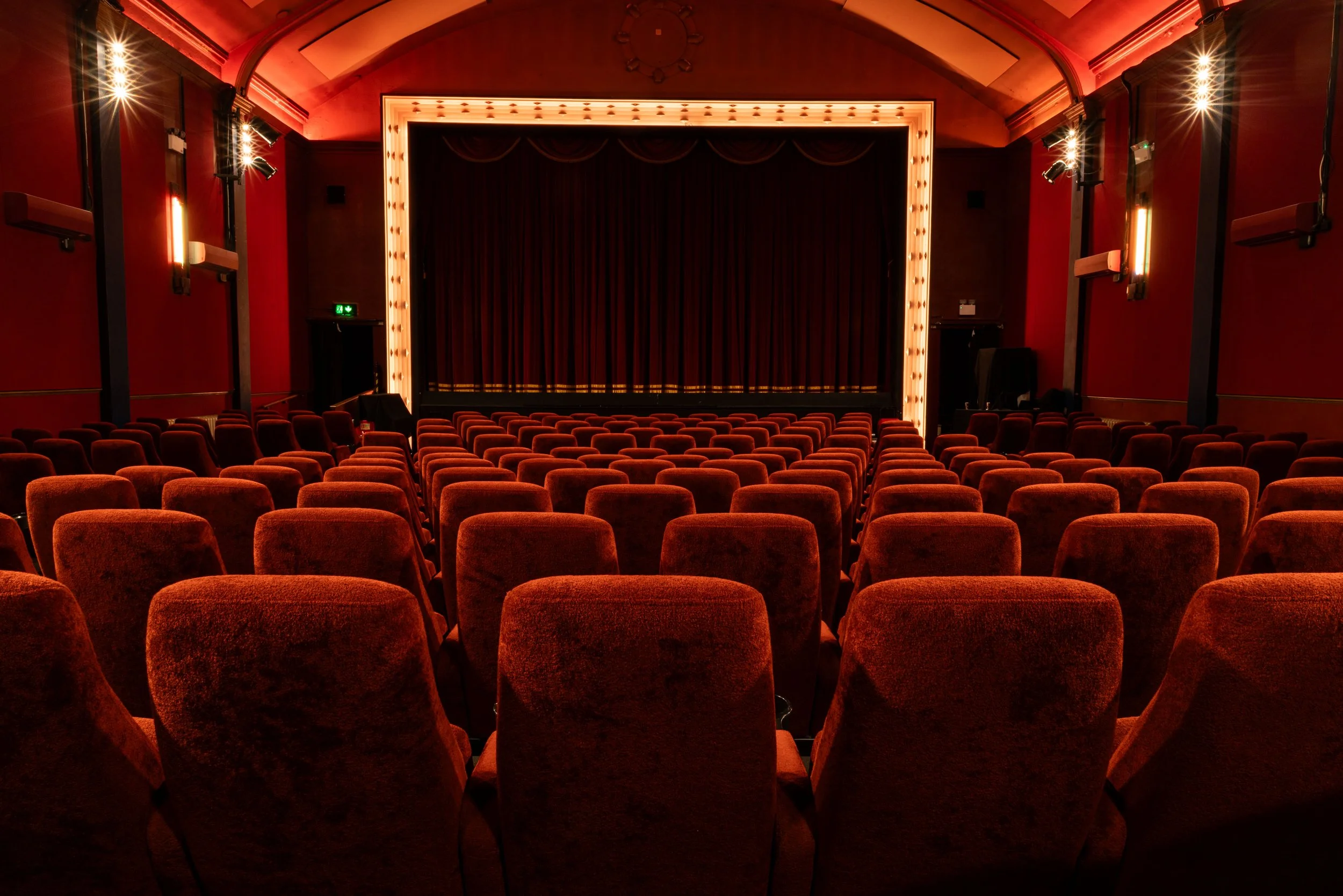 Empty theater with rows of red upholstered seats facing a stage with red curtains. The theater is dimly lit with warm reddish lighting.