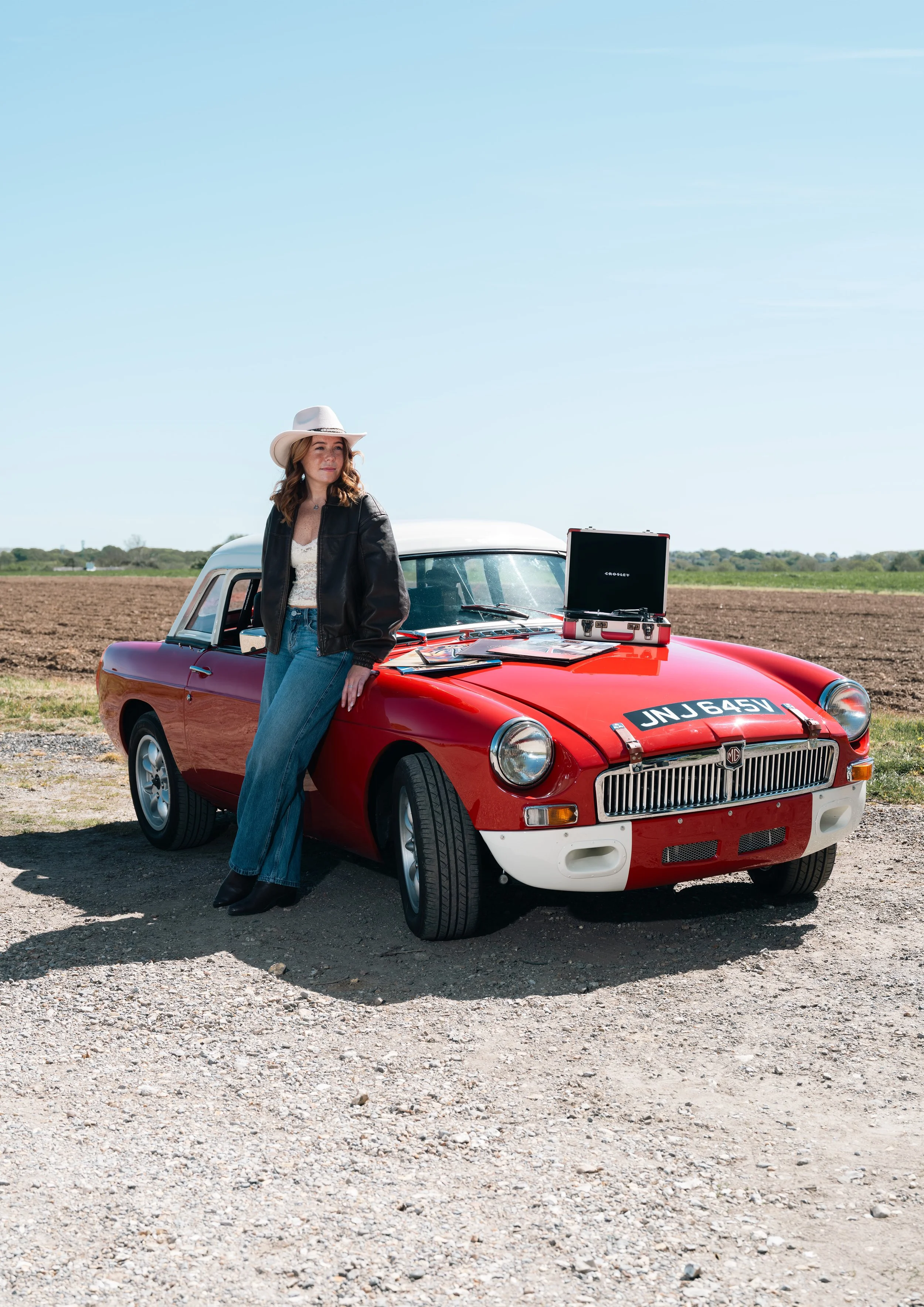 A woman wearing a cowboy hat, black leather jacket, and blue jeans leaning against a red vintage MG convertible car in an open rural area with fields and clear blue sky.