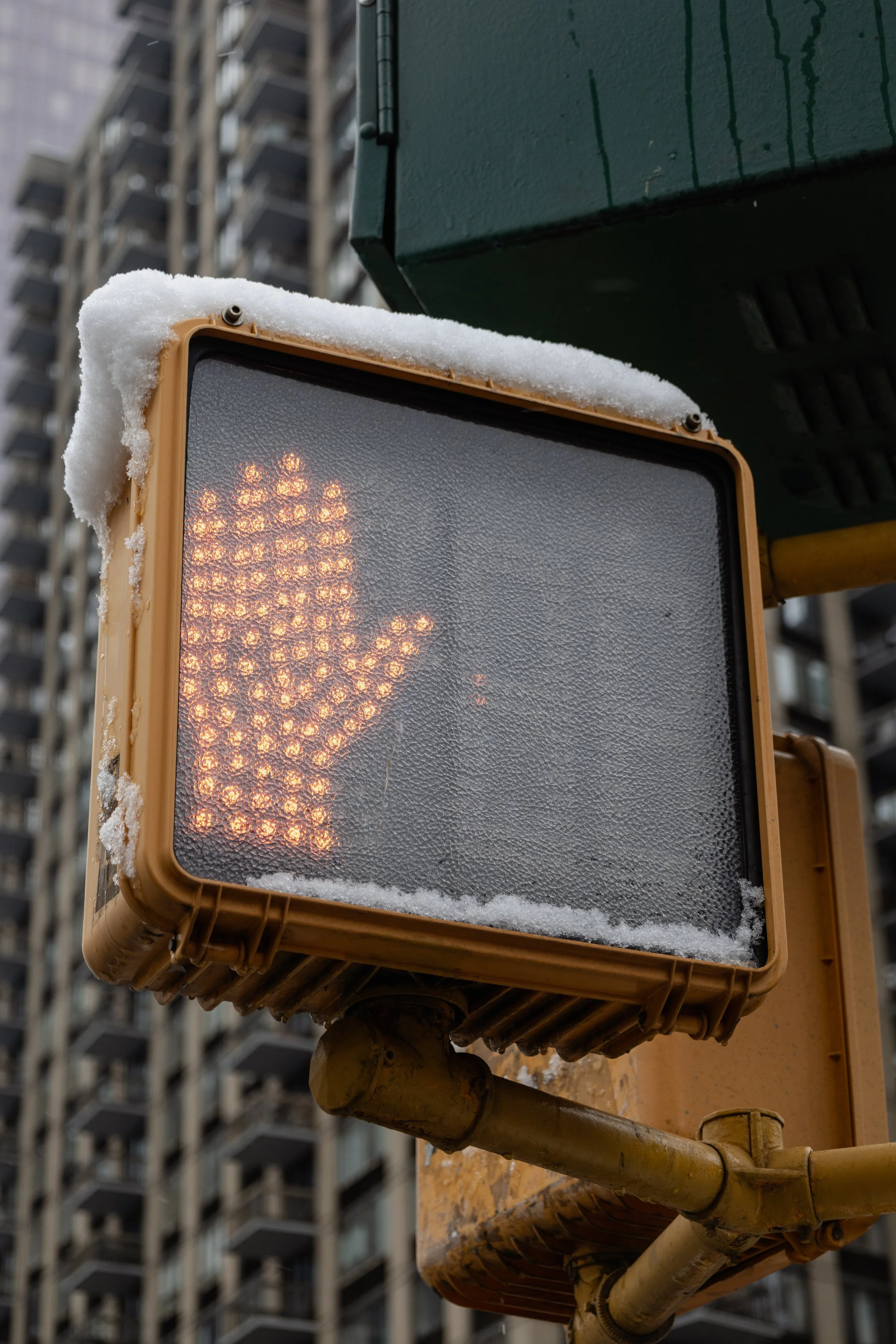 A pedestrian crossing signal with an illuminated hand symbol, indicating stop, covered with snow, in an urban environment with high-rise buildings in the background.