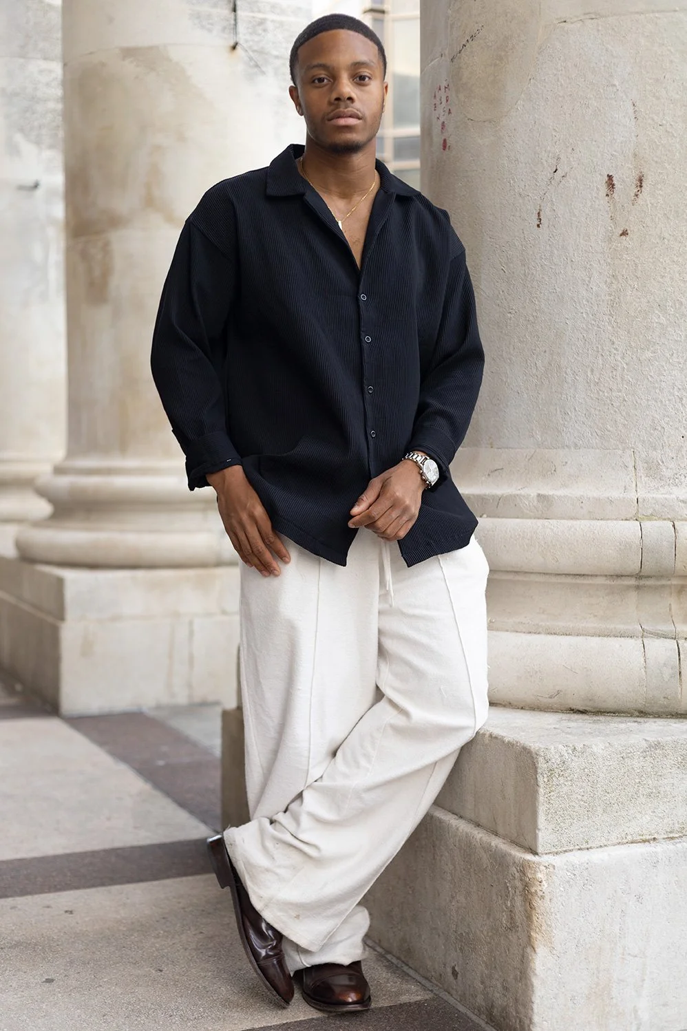 A young man standing outdoors against stone columns, wearing a black long-sleeve shirt, white loose-fitting pants, brown shoes, a watch, and a gold necklace.
