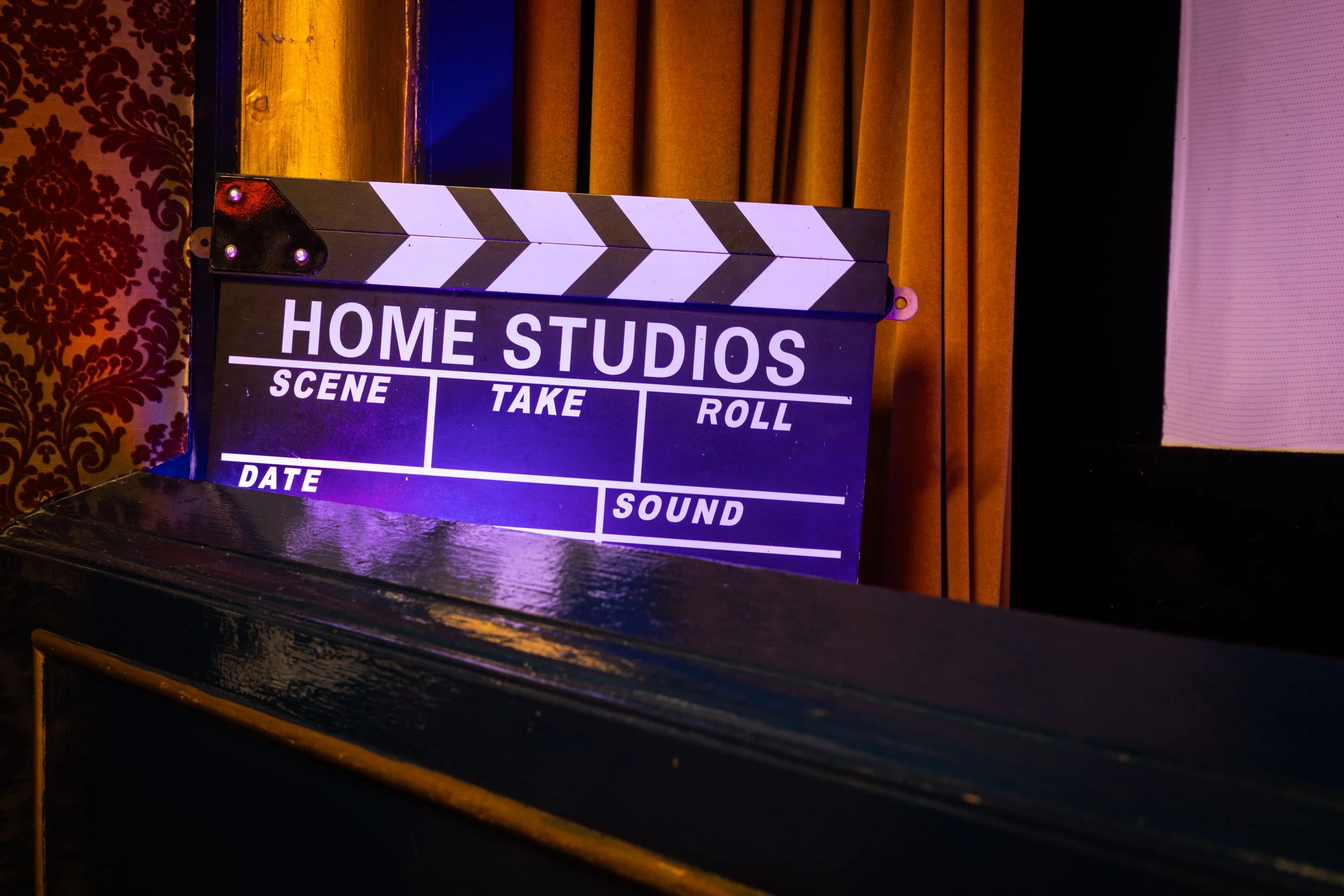 A film clapboard with the words 'Home Studios' in white letters on it, positioned on a dark surface in front of a background featuring gold curtains and decorated wallpaper.