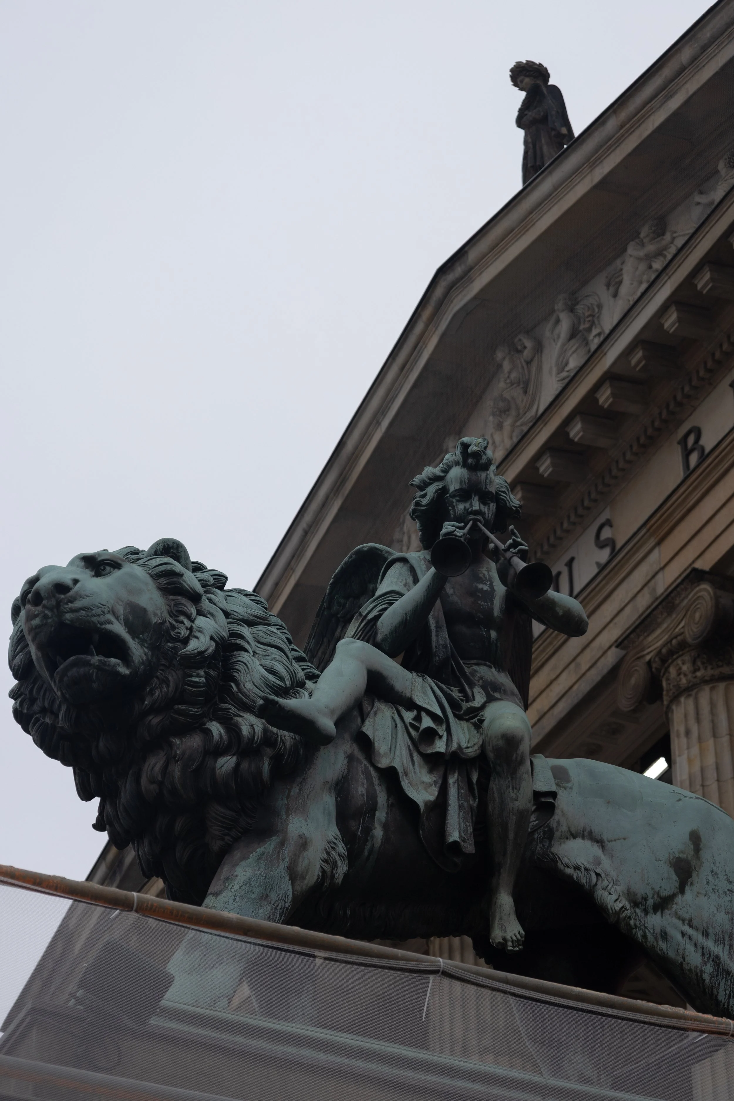 Bronze statue of a lion with a muscular form and flowing mane in front of a building with classical columns and decorative sculptures, with a figure standing on the building's edge in the background.