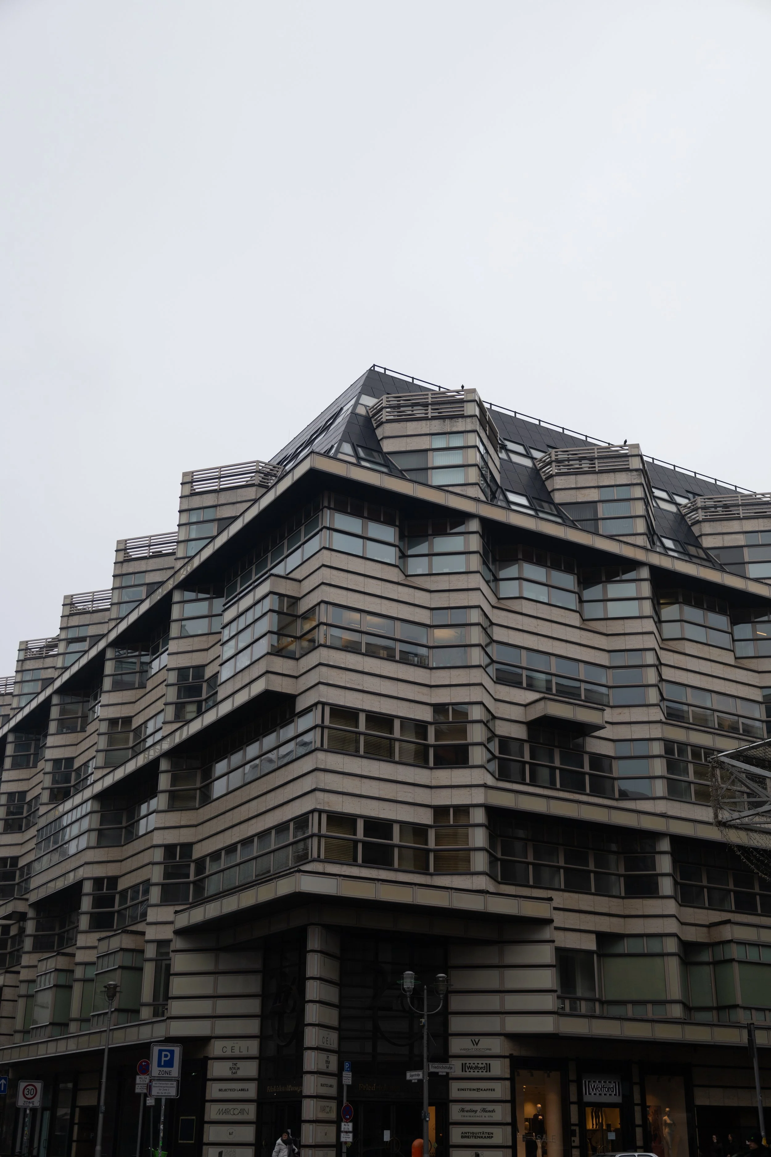 A tall, modern building with multiple balconies and large glass windows in an urban area, with shops on the ground floor and a gray, overcast sky above.