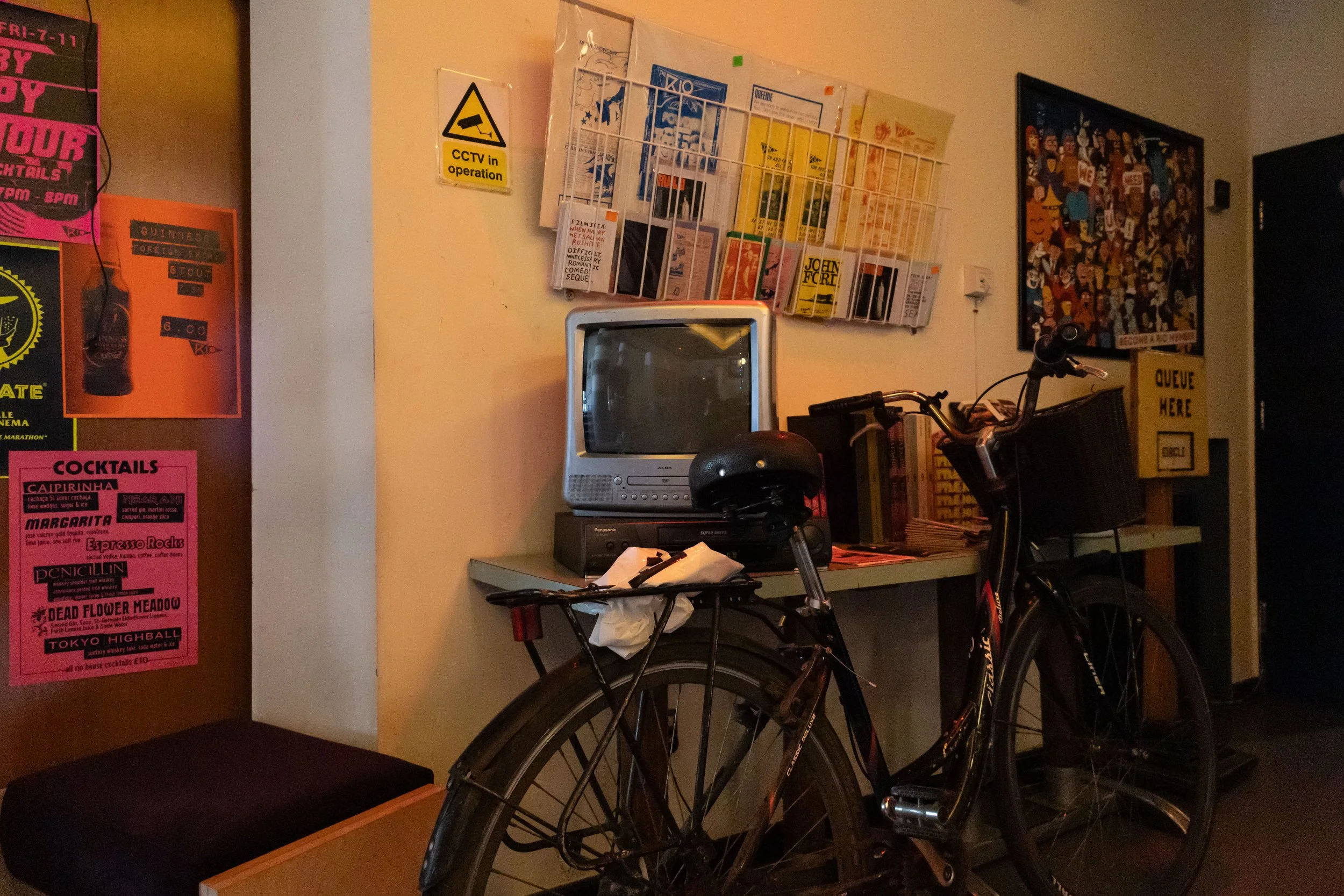 A scene inside a room with posters on the wall, a small TV on a desk, a bicycle parked in front of the table, and a stack of magazines or papers. There is also a yellow newspaper stand with 'QUEUE HERE' written on it, and a framed collage of various 
