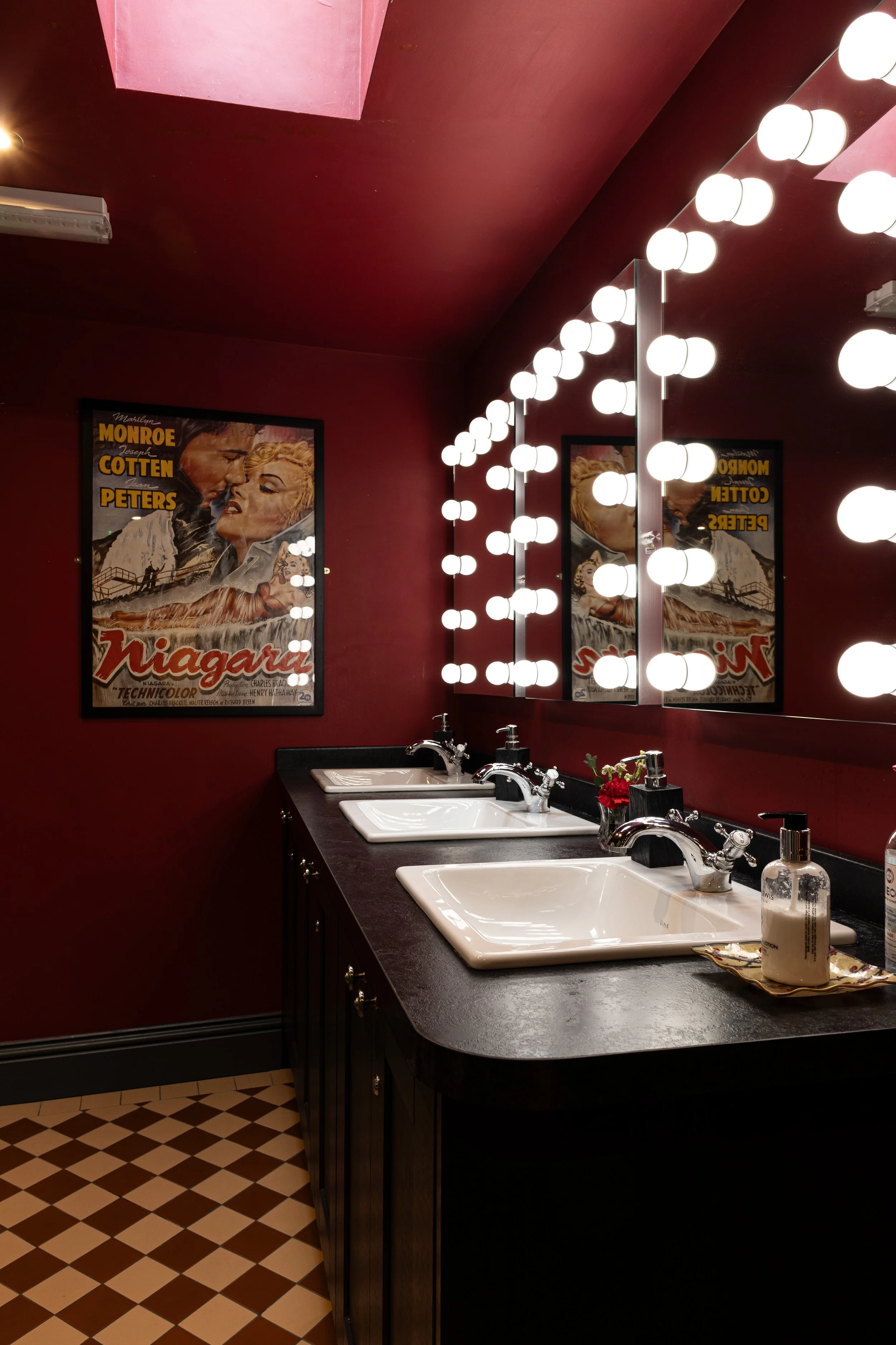 A bathroom vanity with four white sinks and two mirrors surrounded by round Hollywood-style lights, dark cabinetry, red walls, a framed vintage movie poster, and a tiled floor.