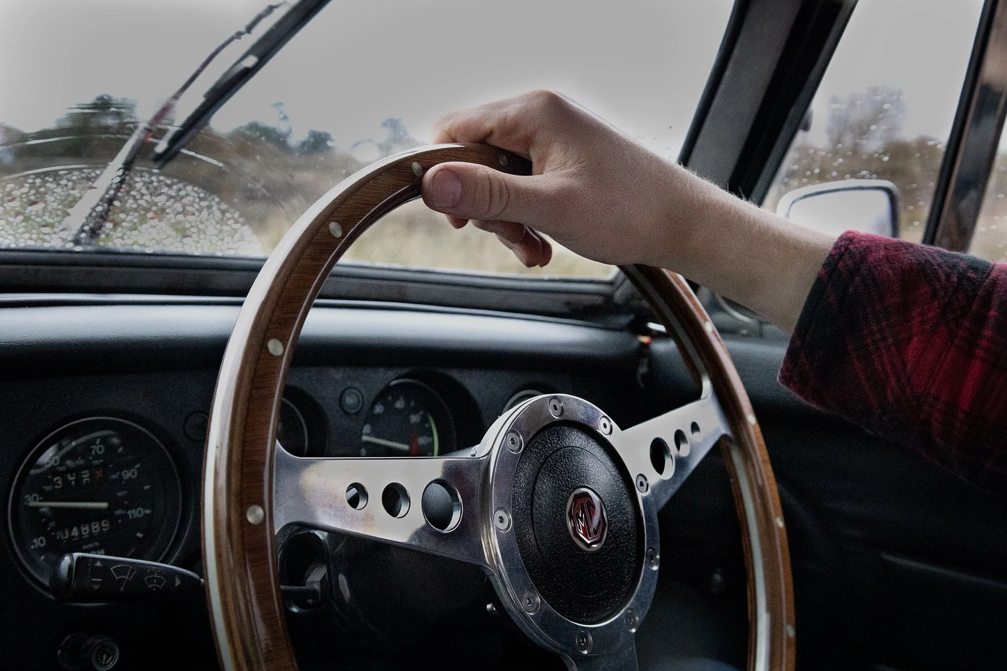 Person holding wooden-rimmed steering wheel inside a vehicle, with raindrops on the windshield and a blurry outdoor scene in the background.