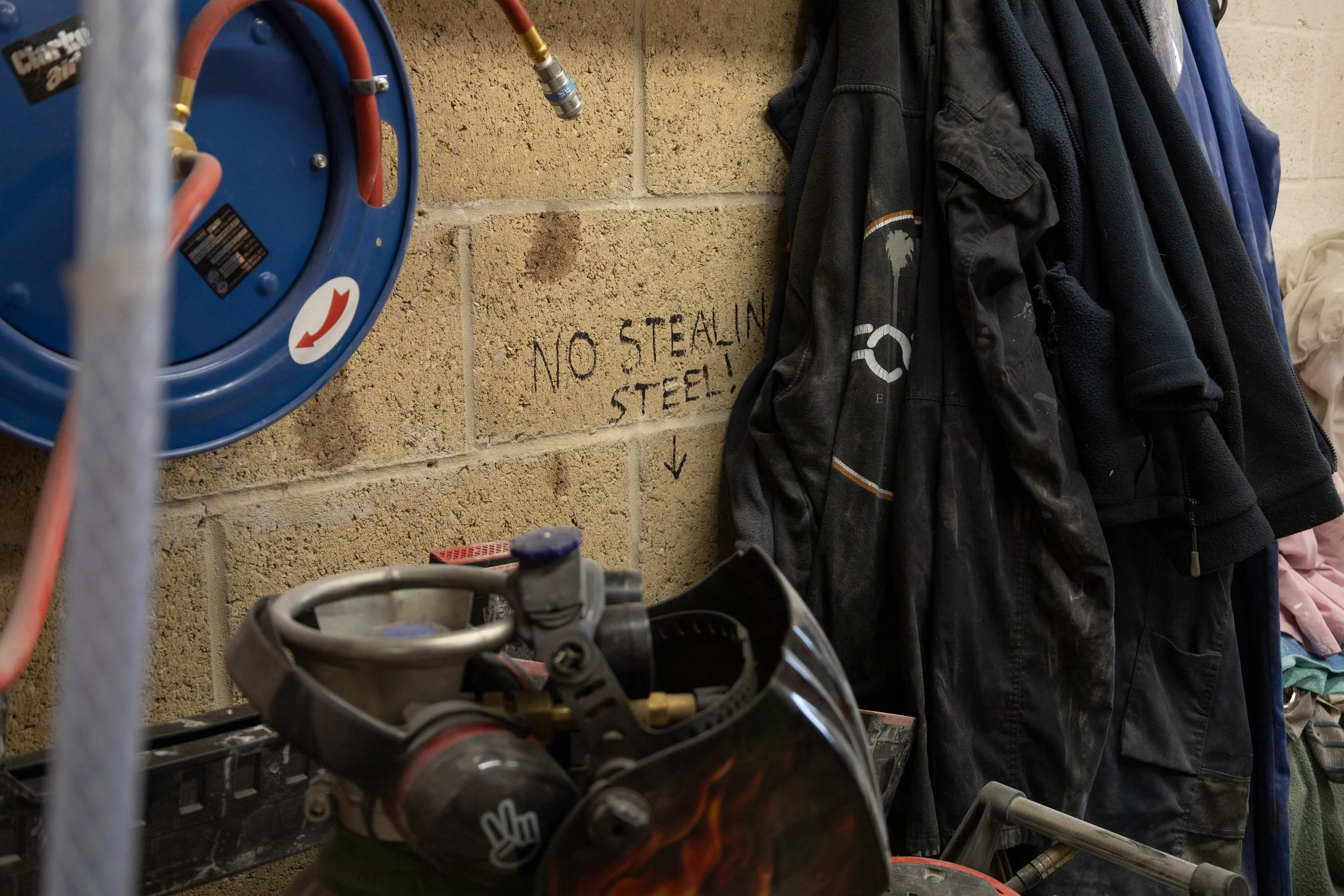 A garage or workshop space with garage jackets hanging on the wall and a sign that says 'NO STEALING STEEL!' in black paint. Part of a motorcycle handlebar is visible at the bottom of the image.