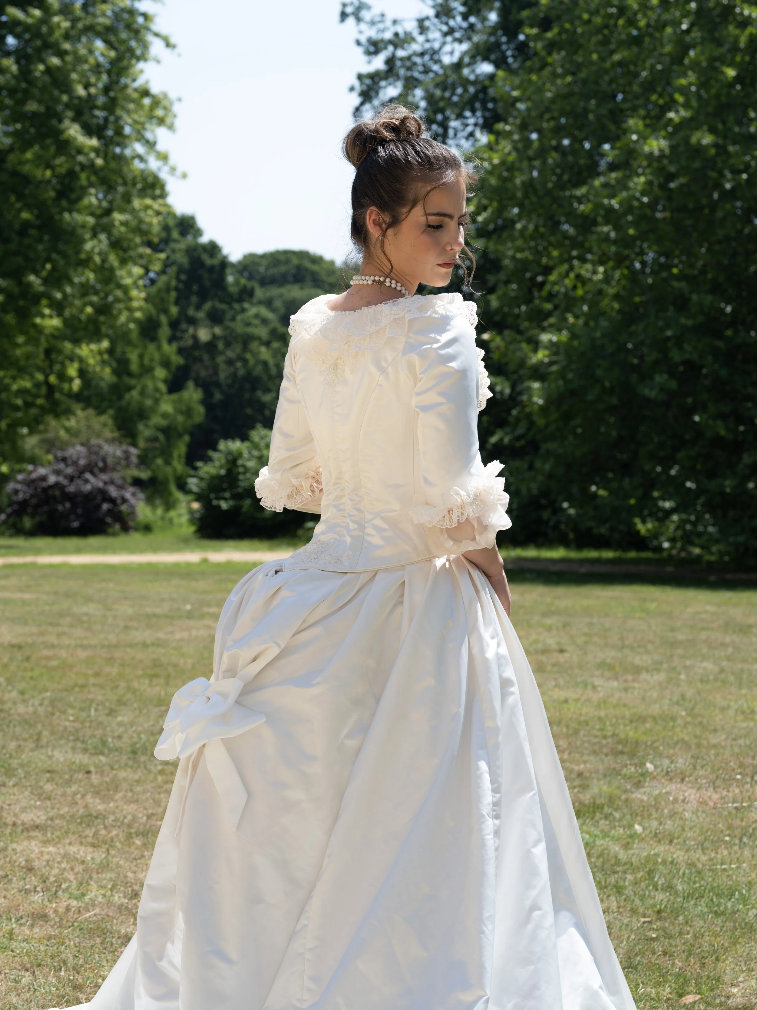 A young woman in a vintage white dress with lace details and puffy sleeves outdoors during daytime, with a background of green trees and grass.