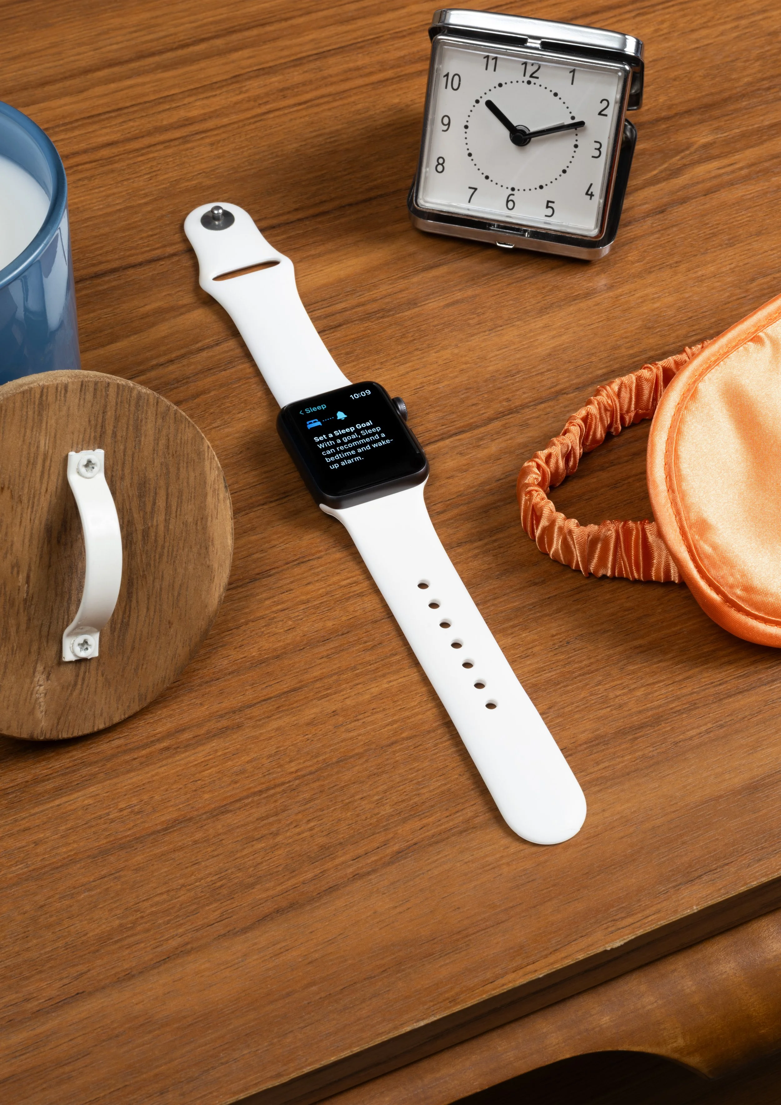 A wooden desk with a blue mug of milk, a white sleep tracker, a foldable silver clock showing around 10:09, an orange sleep mask, and a wooden coaster.