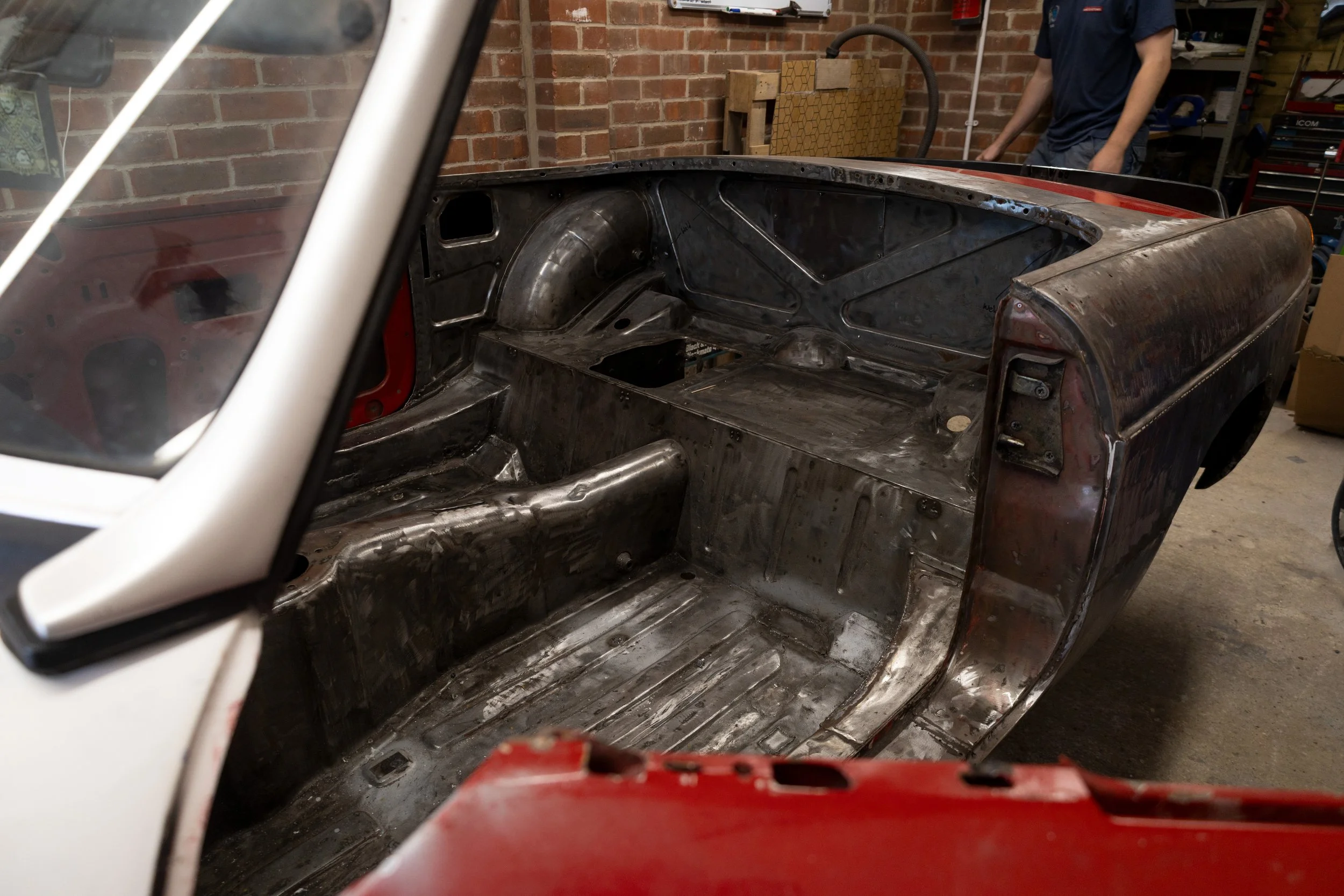Inside a car's stripped-down metal chassis, with a brick wall in the background, and two people working on the car in a garage.