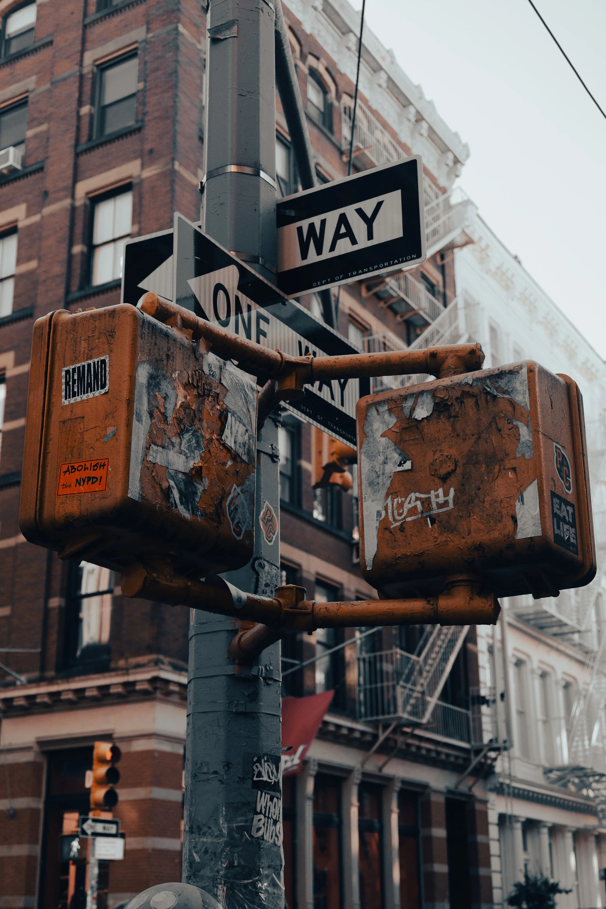 City street corner with a fire hydrant, traffic light, and street signs. Buildings with fire escapes are visible in the background.