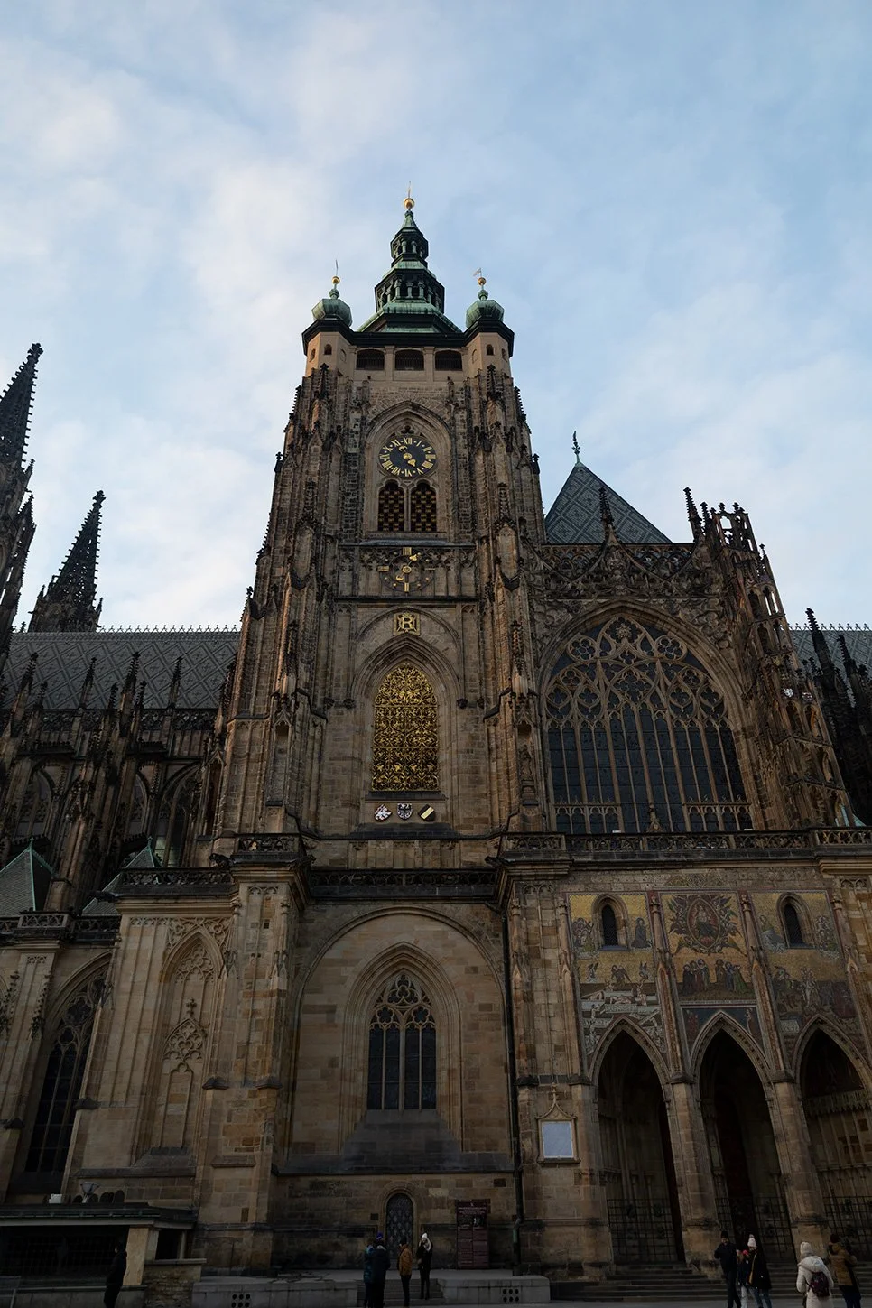 View of a Gothic-style cathedral with a tall clock tower and stained-glass windows, set against a partly cloudy sky.
