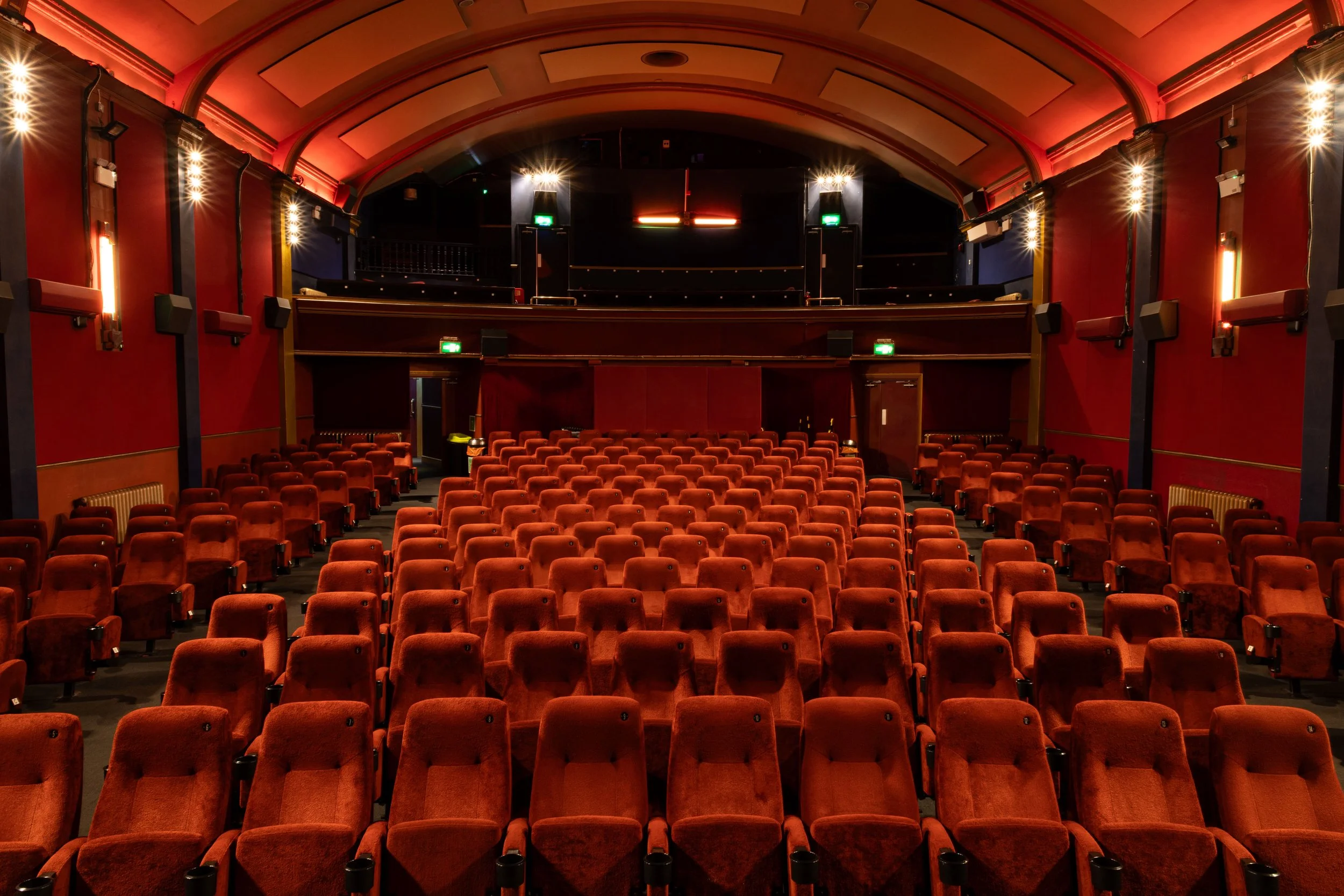 Inside a theater with rows of red velvet seats, a stage with a black backdrop, and warm lighting creating a cozy atmosphere.