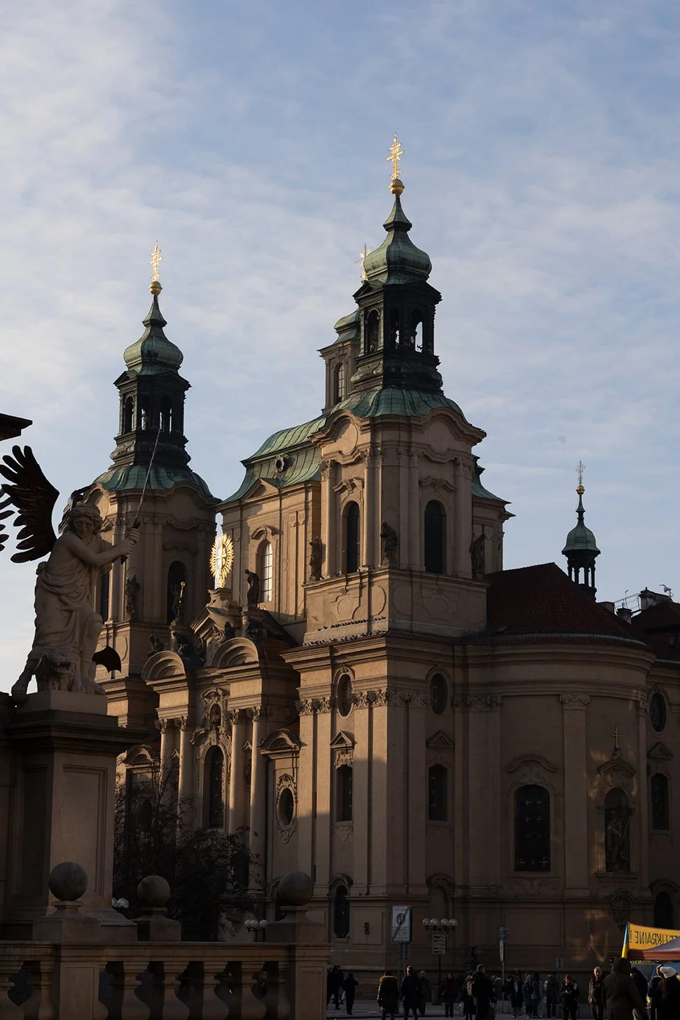 A historic baroque church with ornate architecture and spires, located in a bustling city square with people walking by and a statue of an angel in the foreground.