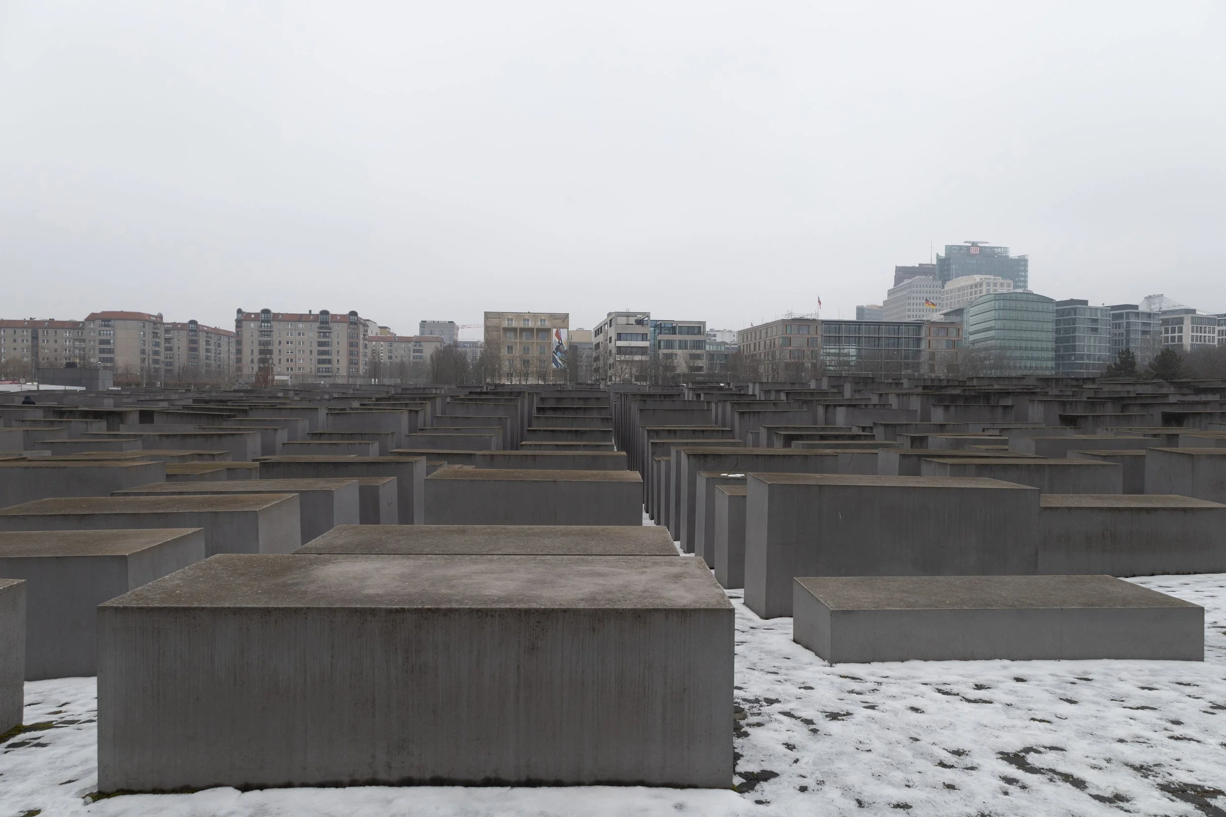 Memorial wall with numerous concrete blocks in a grid pattern in front of a city skyline under a cloudy sky on a cold winter day.