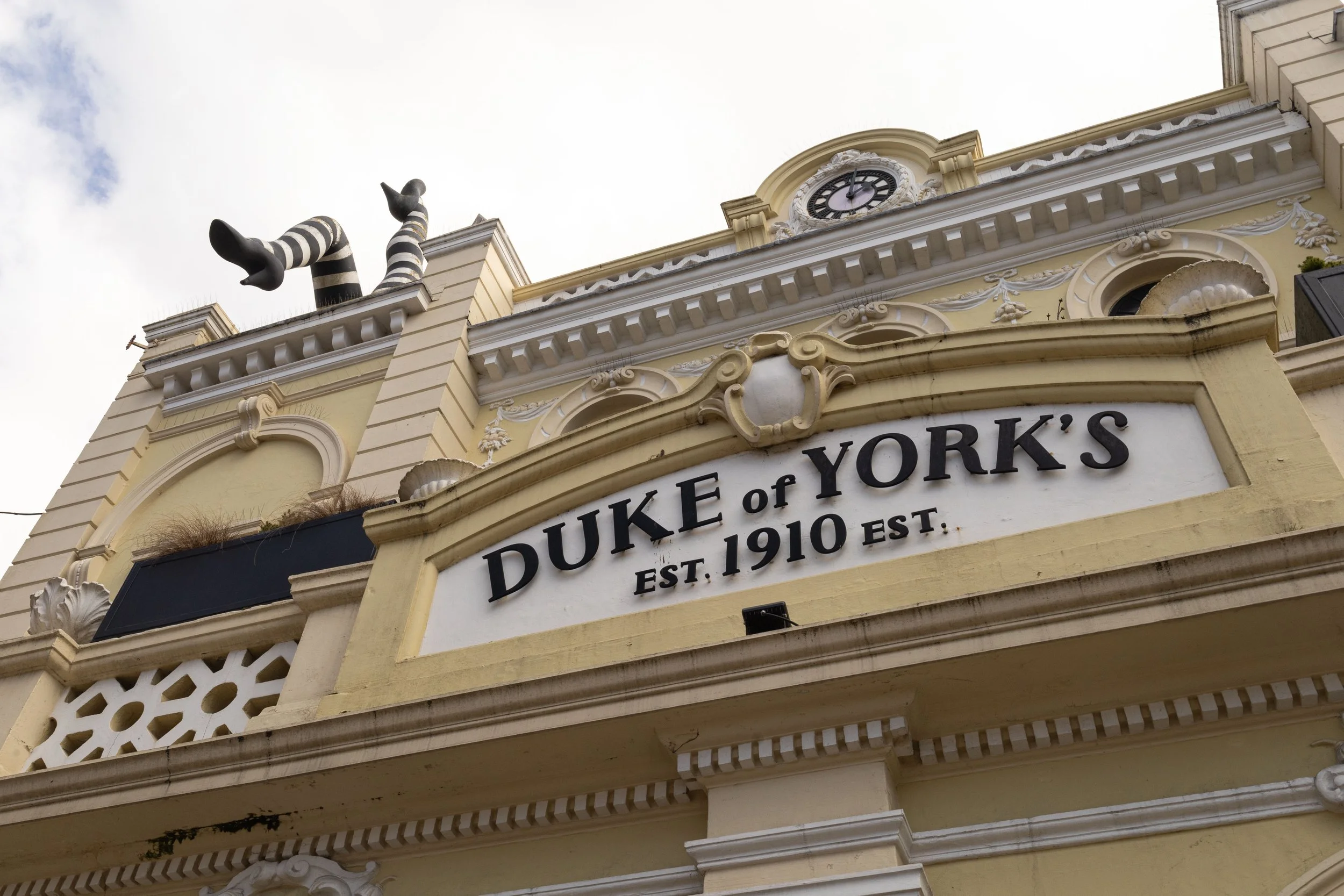 The corner of a historic building displaying a clock with black hands and numerals, ornate architectural details, a sign that reads 'DUKE of YORK'S' with 'EST. 1910' beneath it, and a black-and-white striped sculpture on the roof.